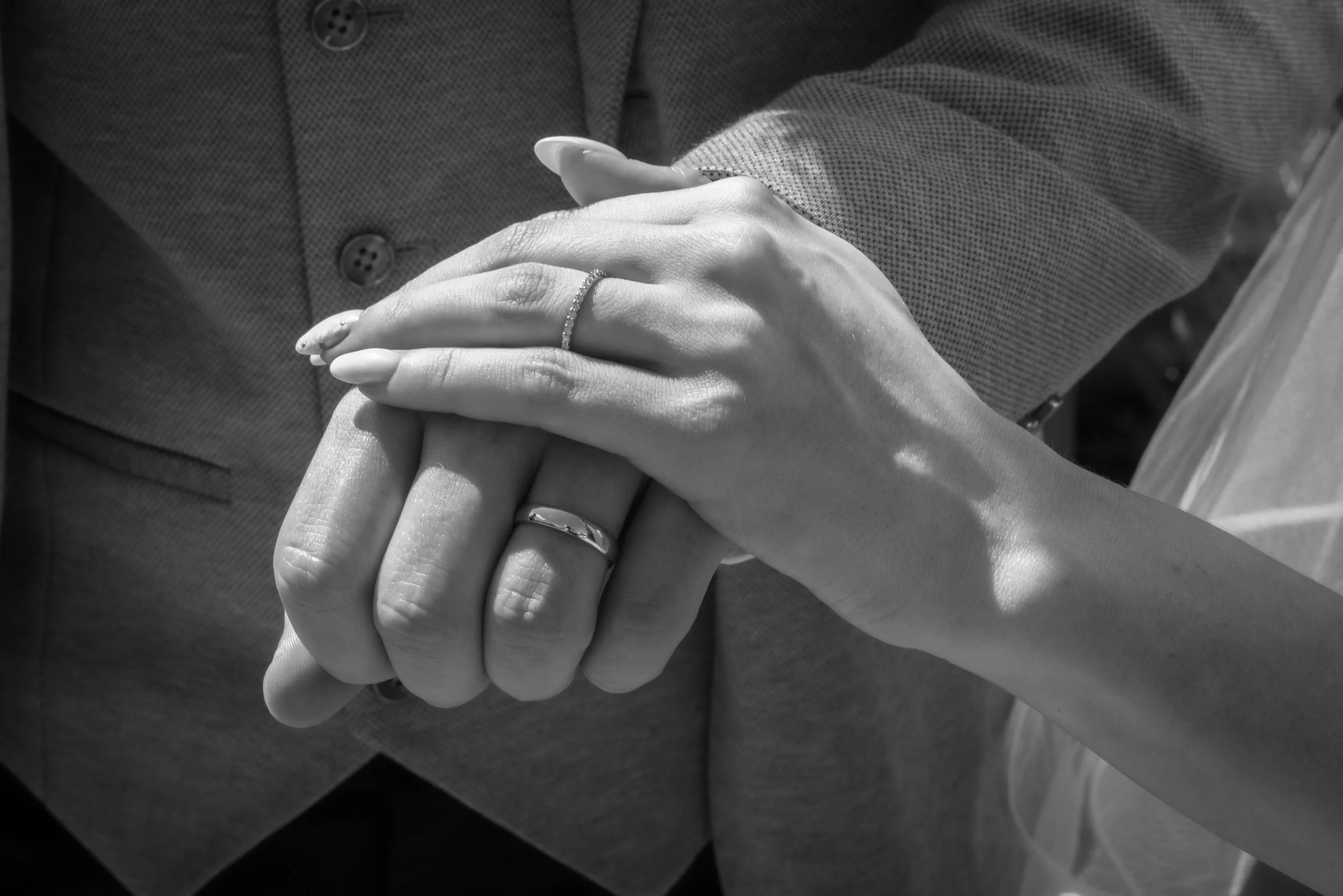 Close-up of a couple holding hands, with visible wedding rings, in black and white. One person is wearing a textured dress shirt.