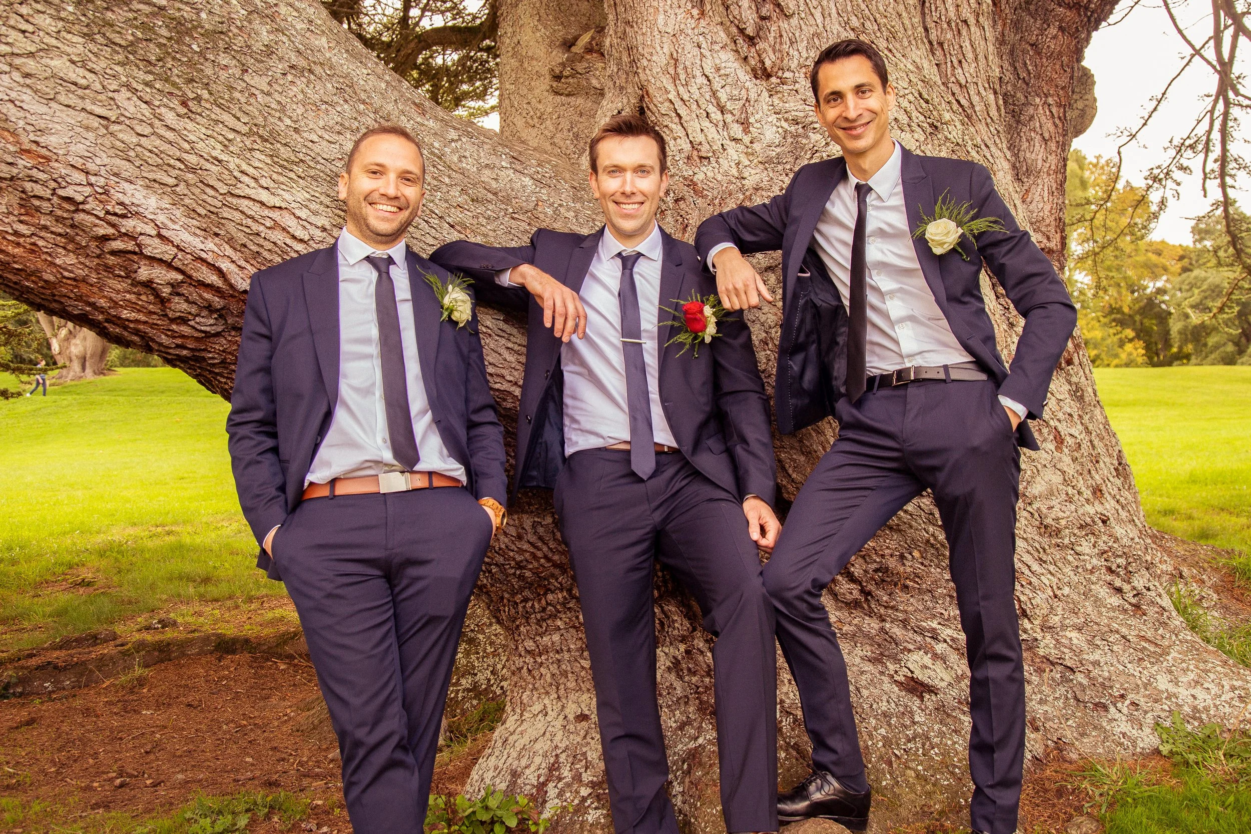 Three men in matching navy business suits with white shirts and ties, each with a boutonniere, standing outdoors by a large tree with rough bark in a park-like setting, smiling at the camera.