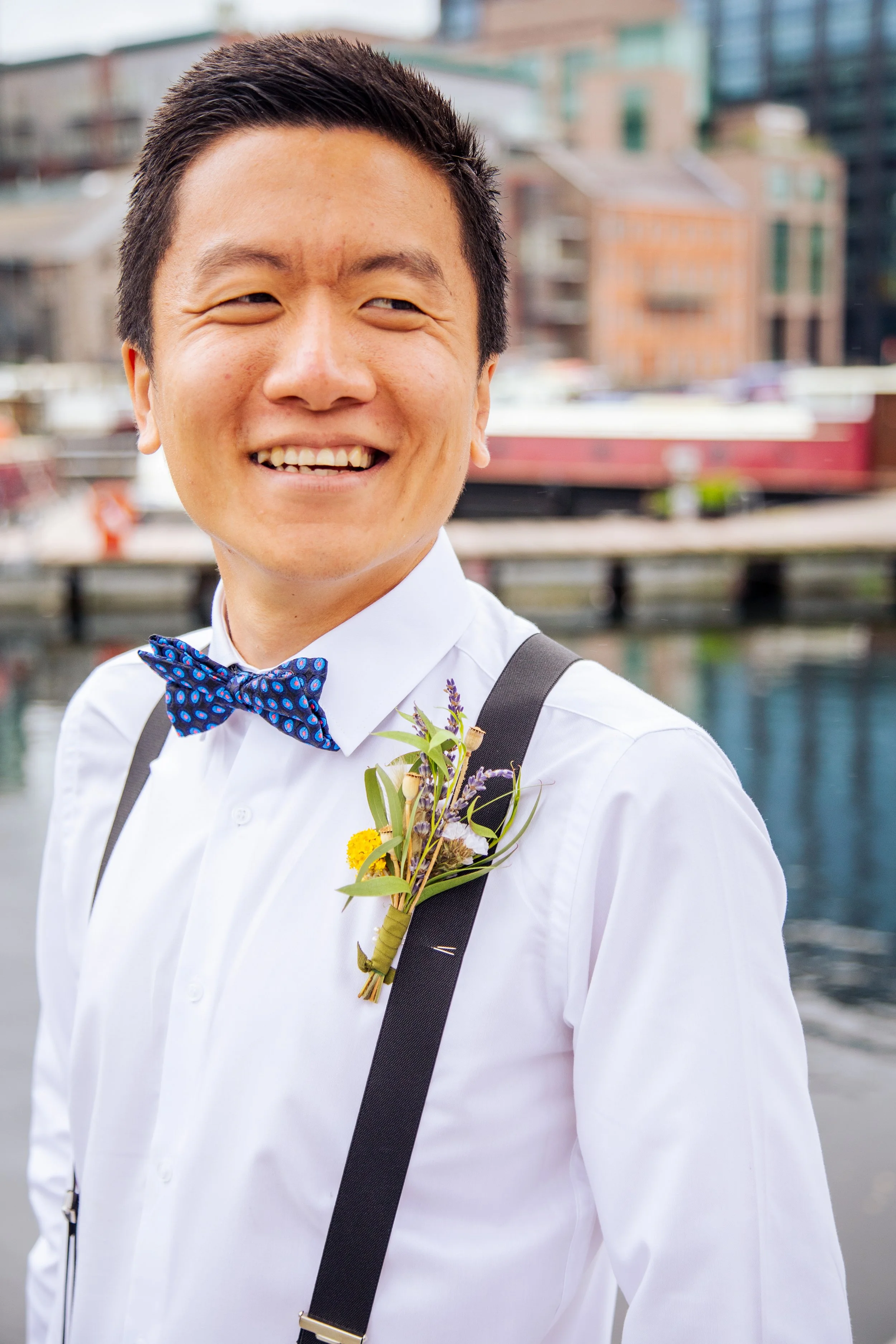 A smiling man in a white shirt with a bow tie, wearing suspenders, standing outdoors with a cityscape background. He has a boutonnière with flowers and greenery pinned to his shirt.