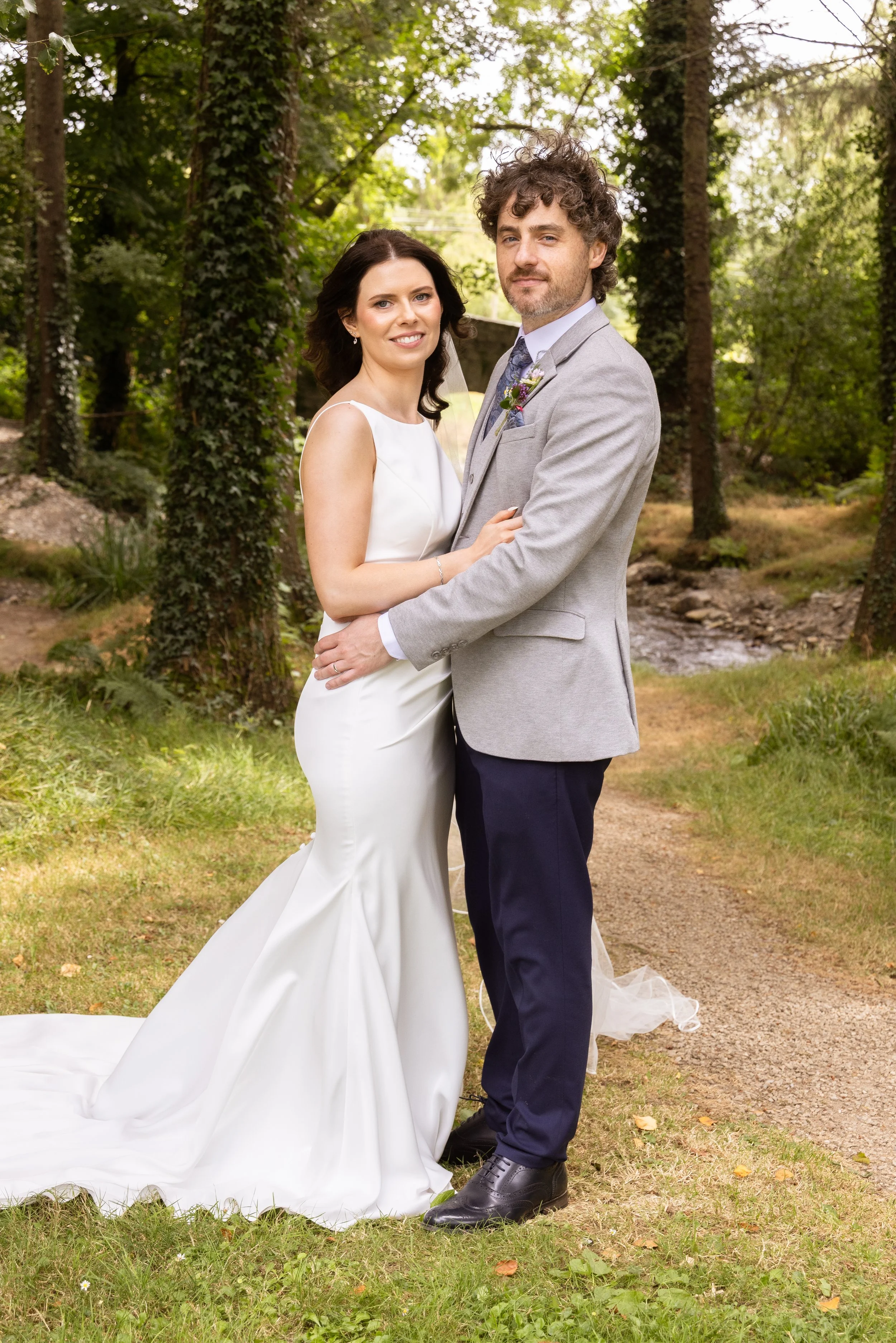A bride and groom standing together outdoors, surrounded by trees and greenery, on their wedding day with the bride in a white gown and the groom in a light gray suit.