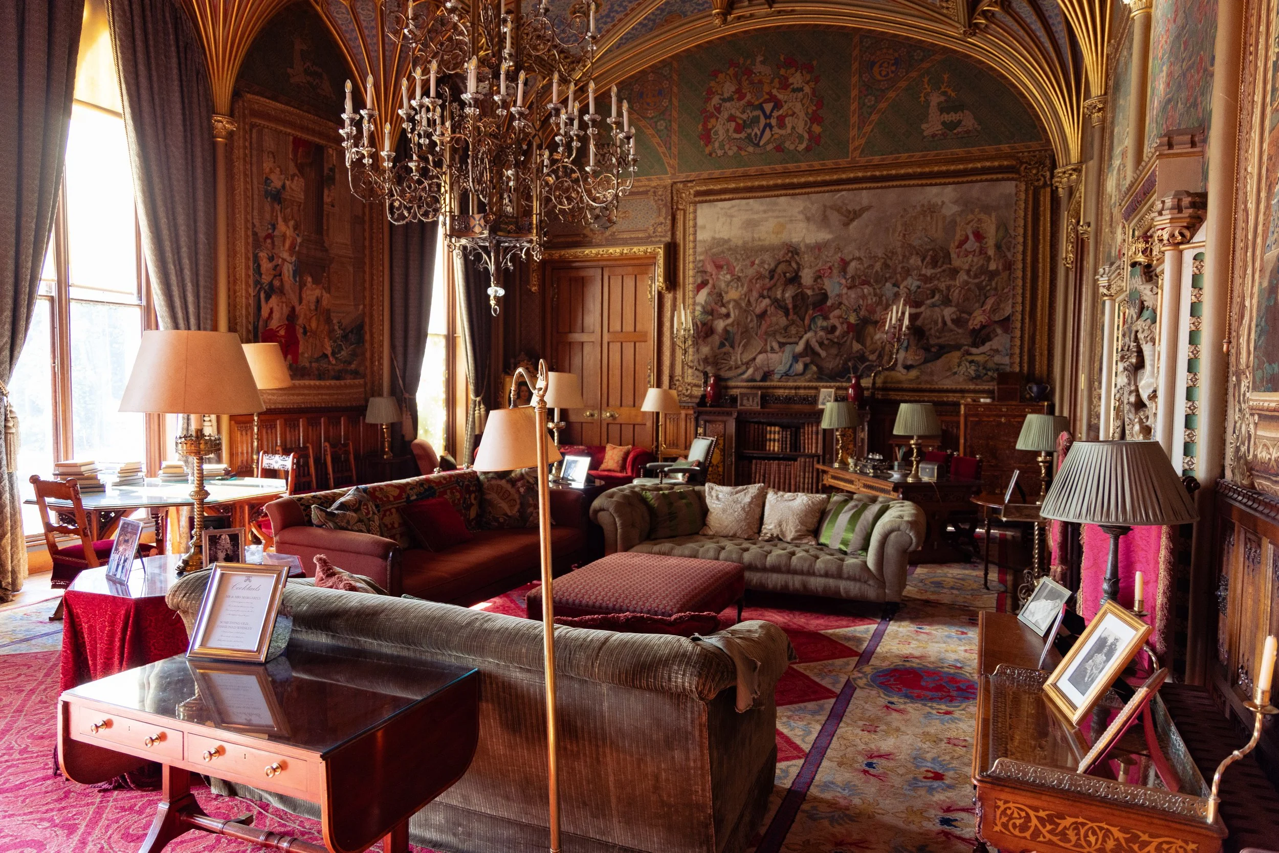 A lavishly decorated room with large windows, luxurious drapes, a chandelier, paintings, and various furniture including sofas, tables, and lamps.