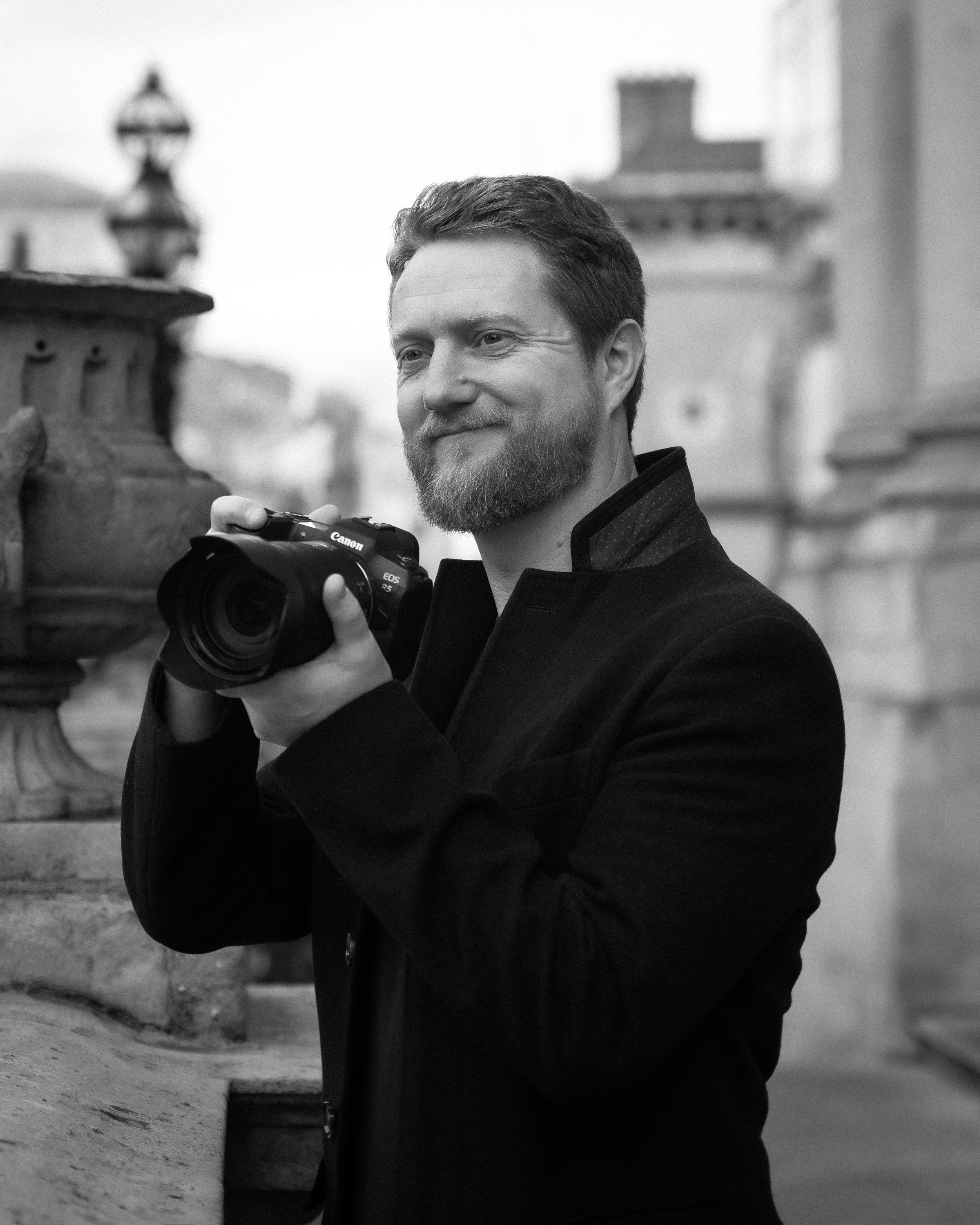 A man with a beard and short hair taking a photograph with a Canon EOS R5 camera on a city street. In the background, there are blurred buildings and a lamp post.