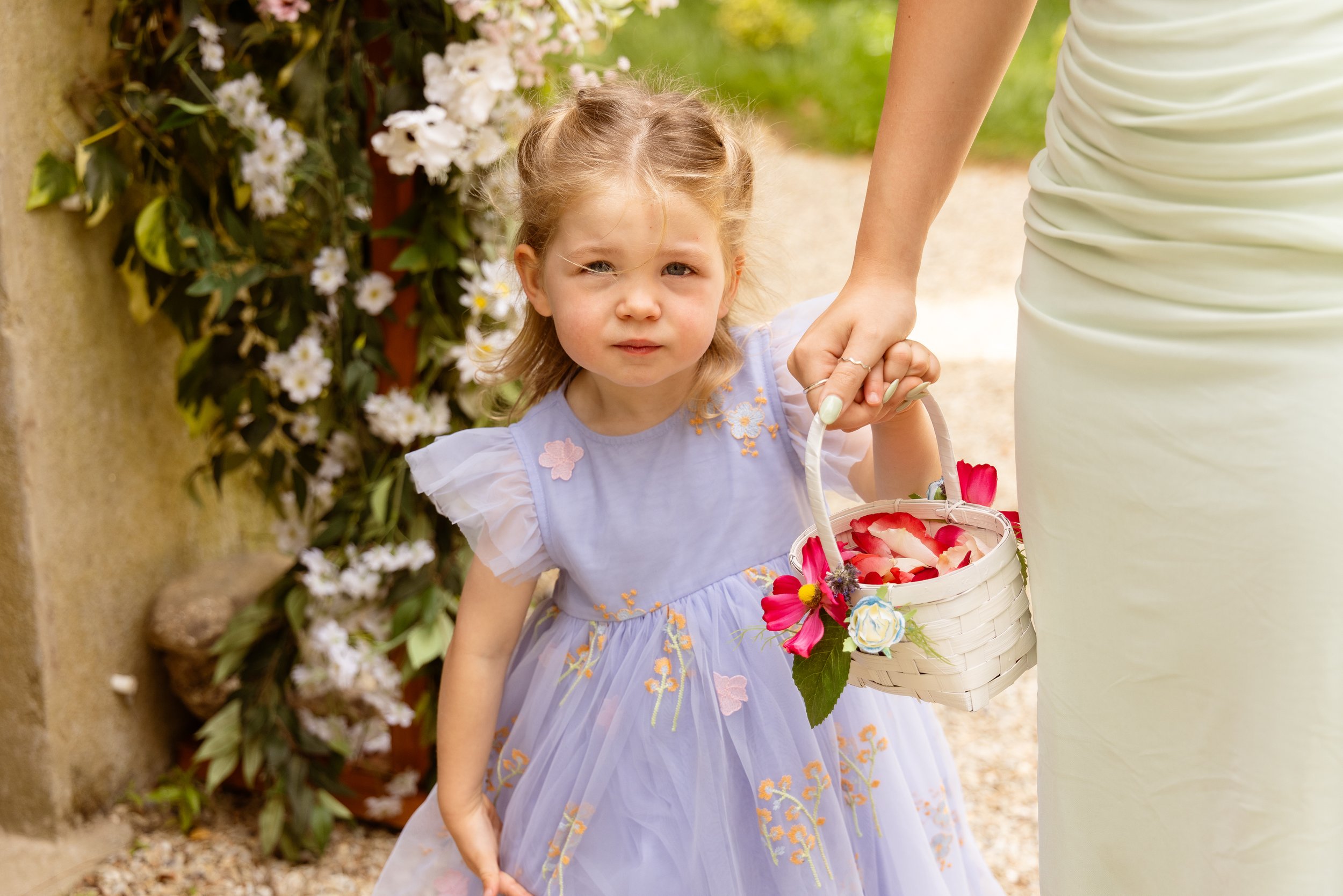 A young girl in a light purple dress with flower embroidery, holding a basket of flower petals after a wedding ceremony, standing outdoors near white flowers and greenery.