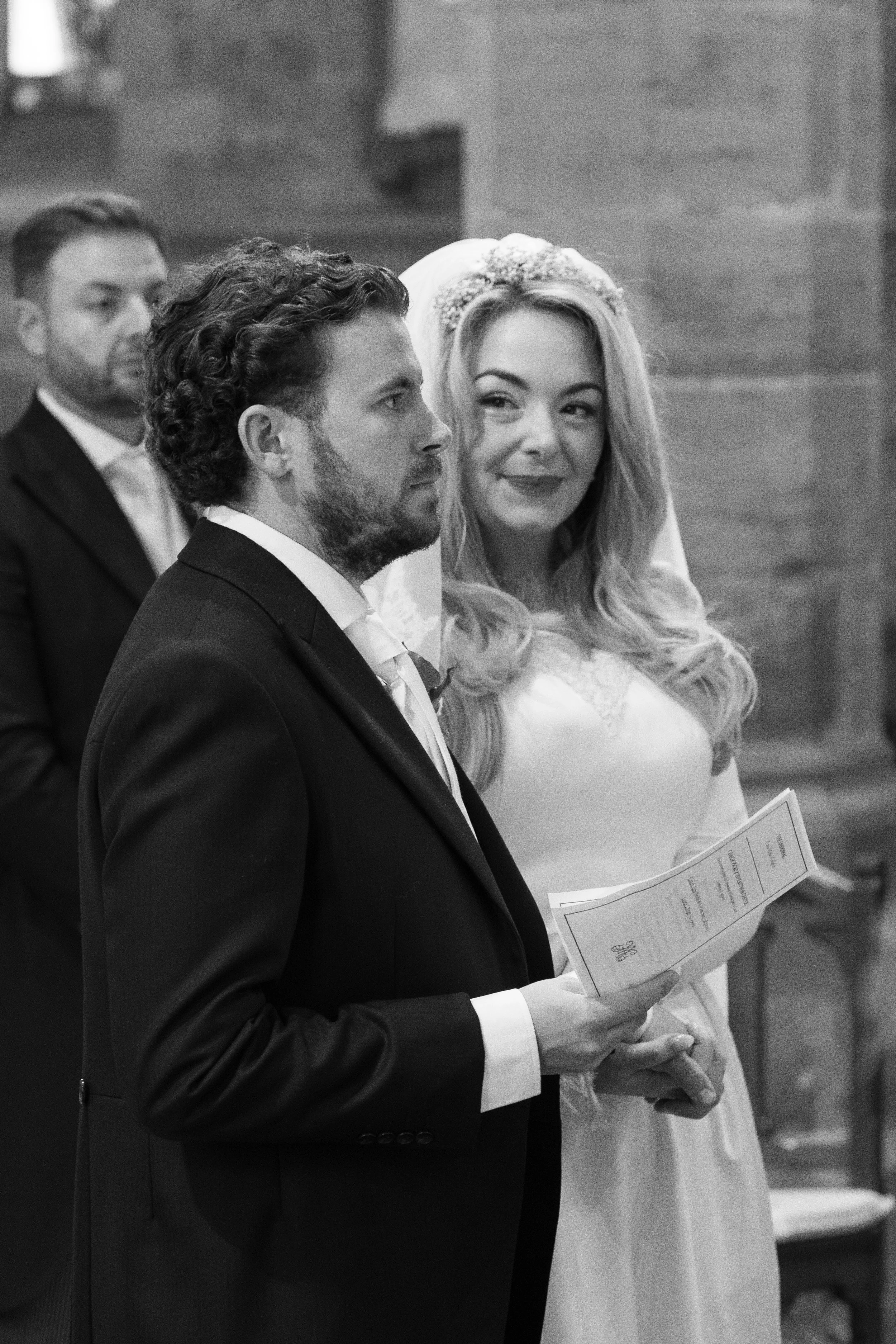 A bride and groom at their wedding ceremony, both holding a program, with the bride wearing a white dress and veil, and the groom in a dark suit, standing in front of a lace curtain with another man in the background.