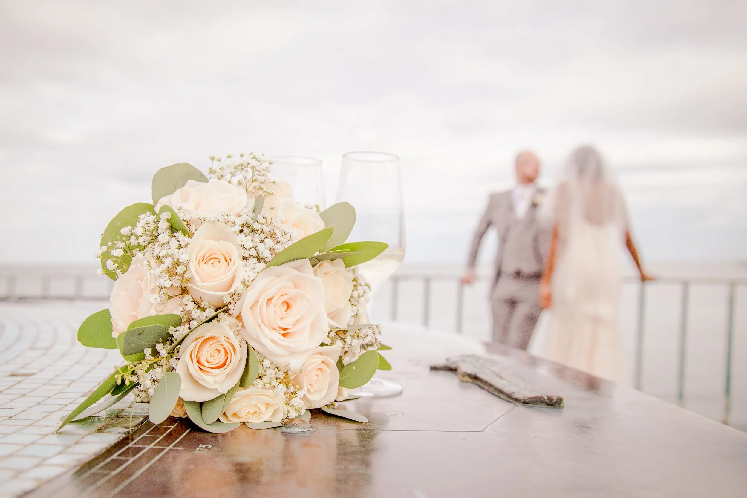 Close-up of a bouquet of pale peach roses, white baby's breath, and greenery on a white table with a wedding couple blurred in the background.