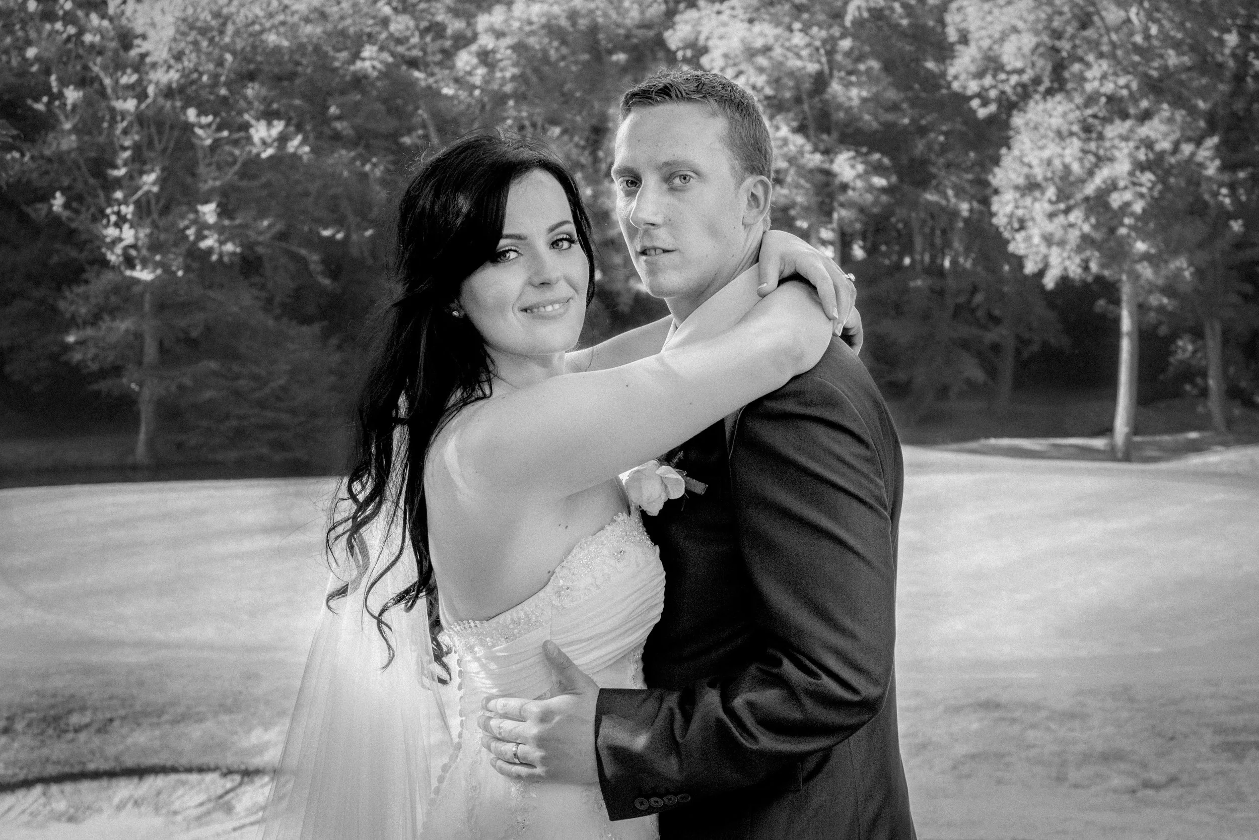 A bride and groom embrace outdoors, with trees in the background, during their wedding.