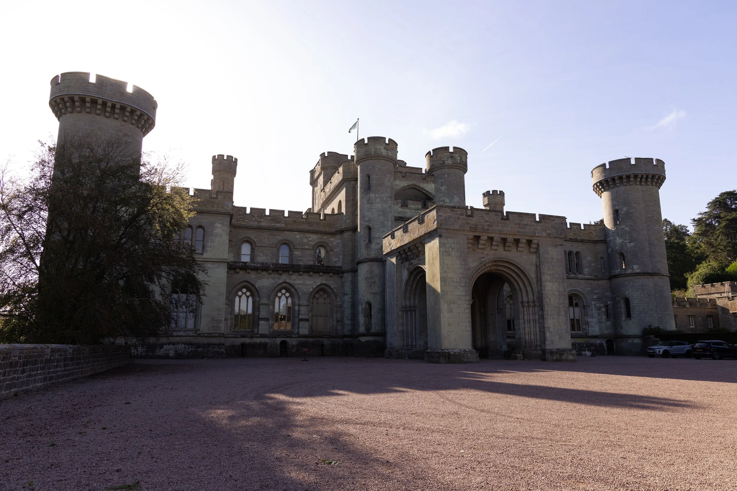 Front view of a historic castle with multiple towers, arched entrance, and stone walls, surrounded by a gravel courtyard and trees, under a clear blue sky.