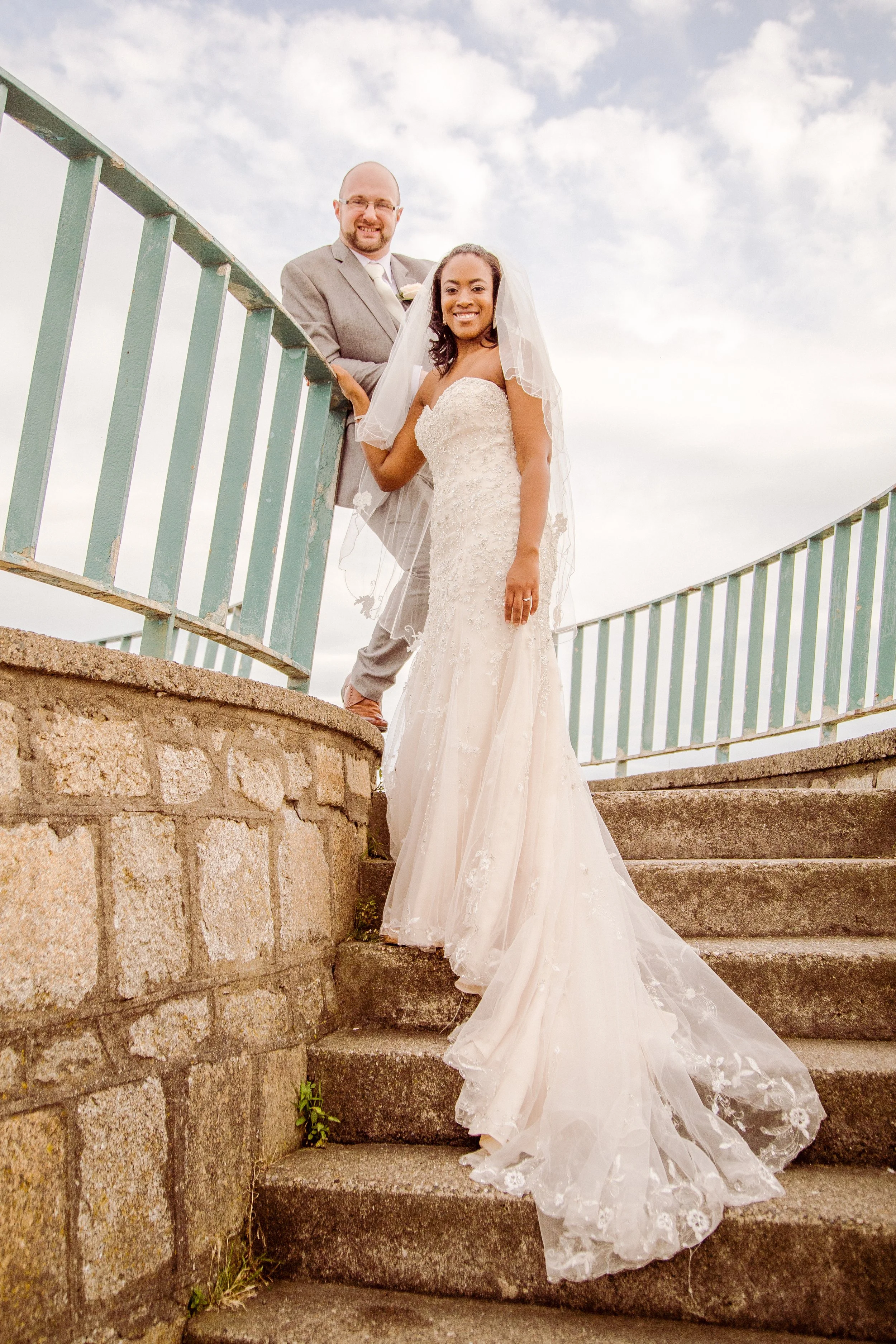 A newly married couple standing on outdoor stone stairs with a blue sky background. The bride is wearing a white lace strapless wedding gown with a long train and veil. The groom is in a grey suit with a white shirt and tie, smiling and holding her h
