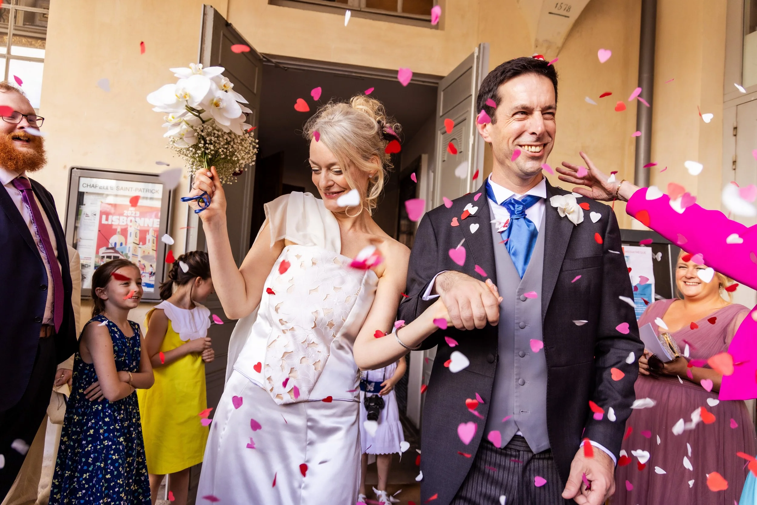 A wedding celebration with people throwing colorful paper hearts in the air. The bride and groom are smiling and holding hands, surrounded by friends and family.