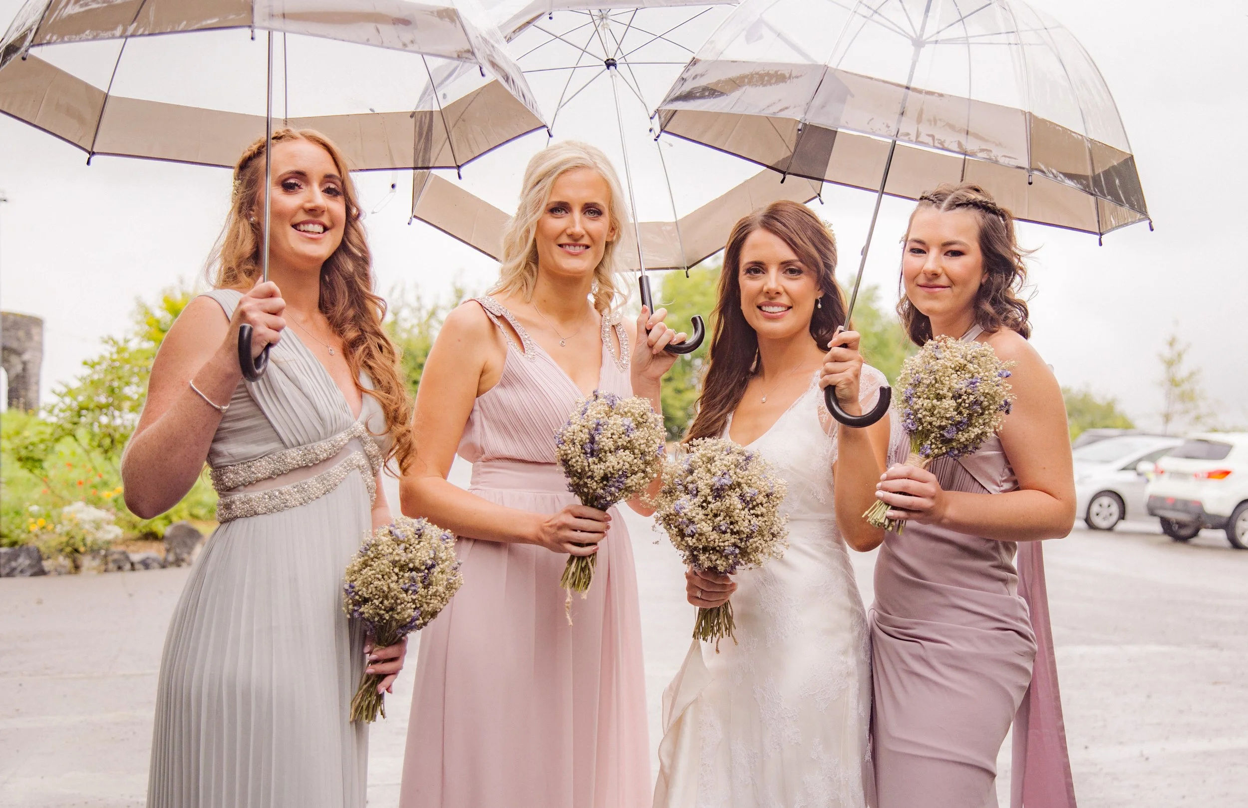 Four women in pastel dresses holding bouquets of flowers, standing under clear umbrellas on a cloudy day.