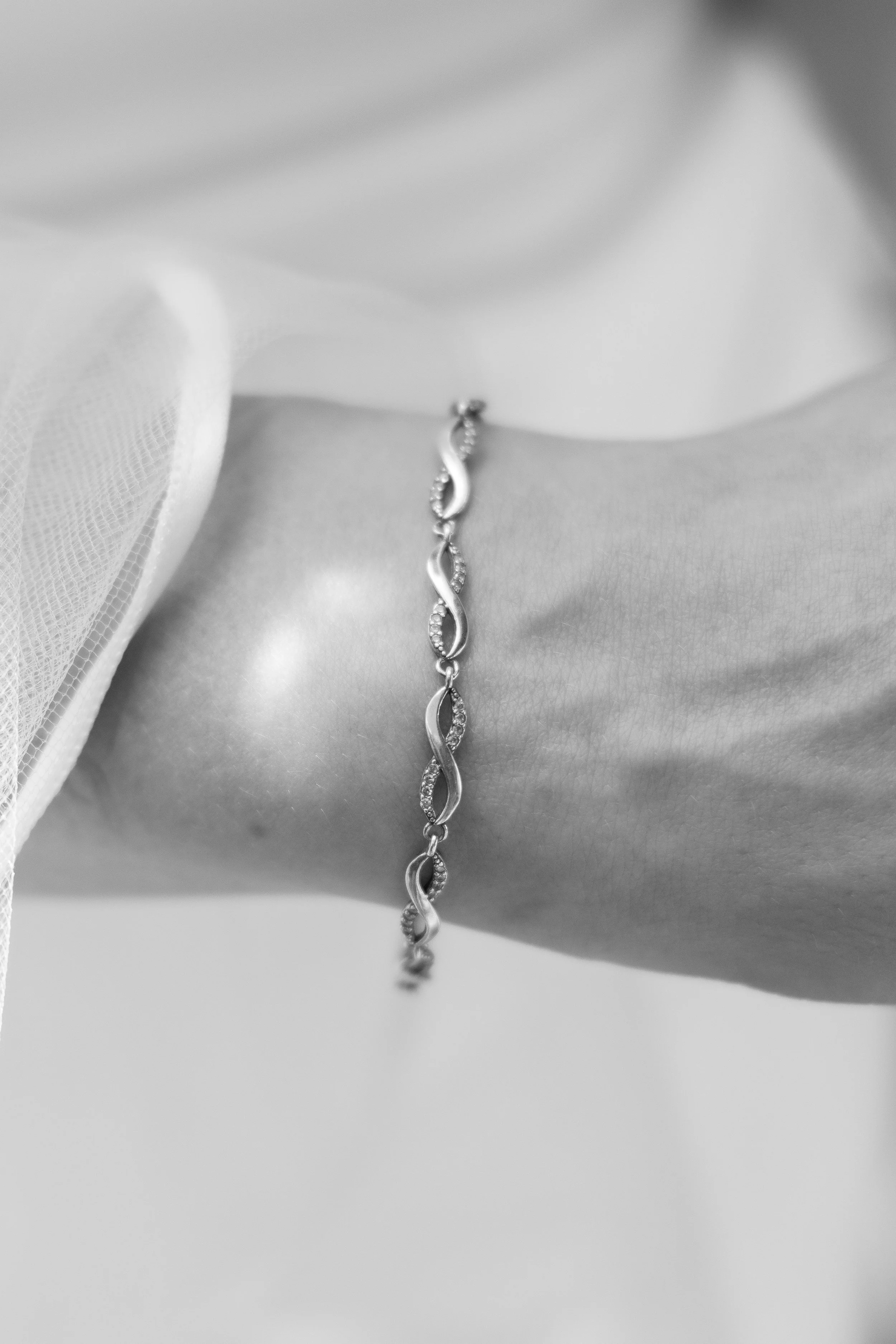 Close-up of a silver necklace with an infinity symbol design encrusted with small gemstones, worn on a person's neck with a white fabric garment.