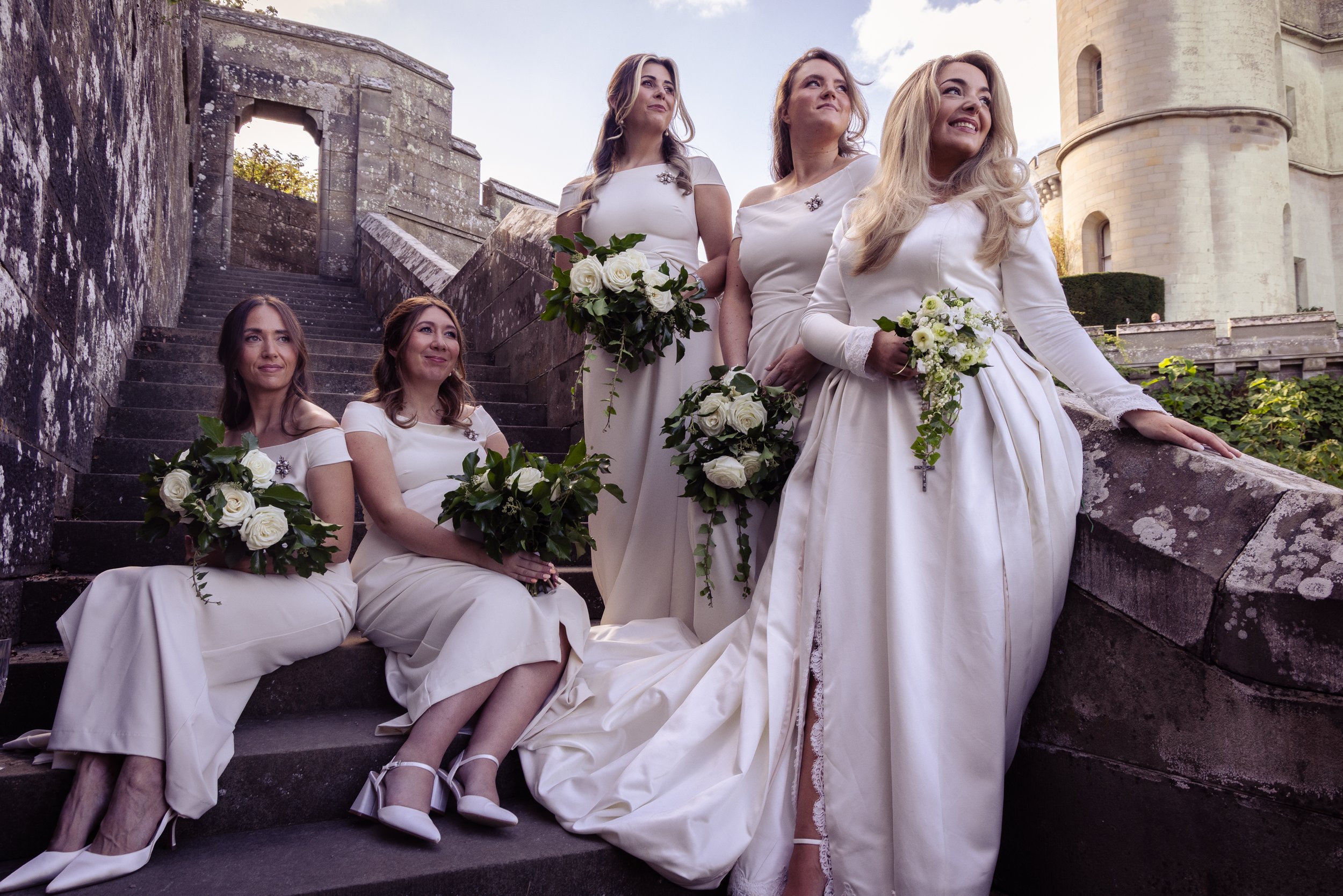 Six women in white dresses holding bouquets of white roses and greenery, posing on outdoor stone steps near a castle, with one women standing and the other five sitting or kneeling.
