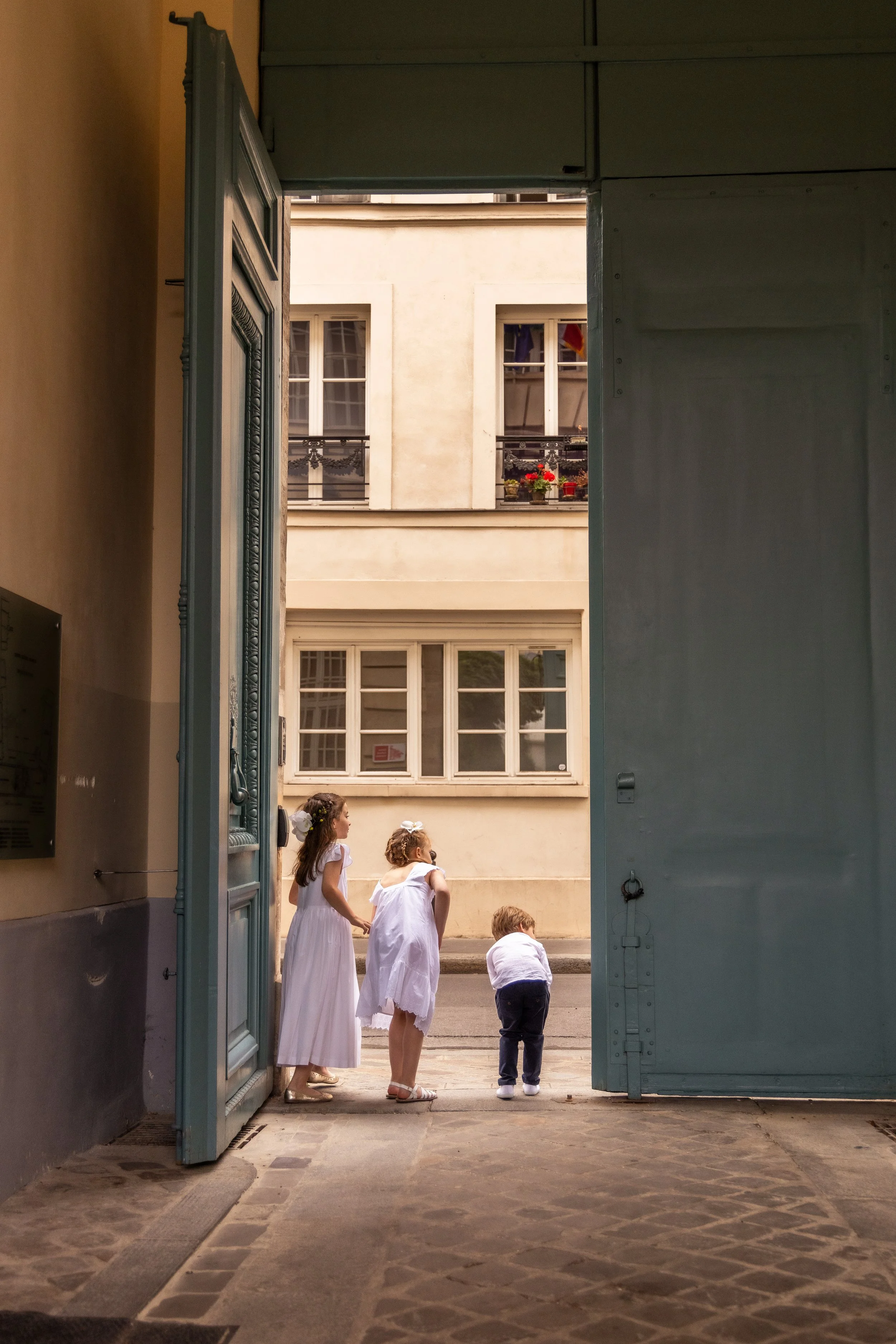 A group of children dressed in white, standing and sitting outside an open door, with buildings and windows in the background.