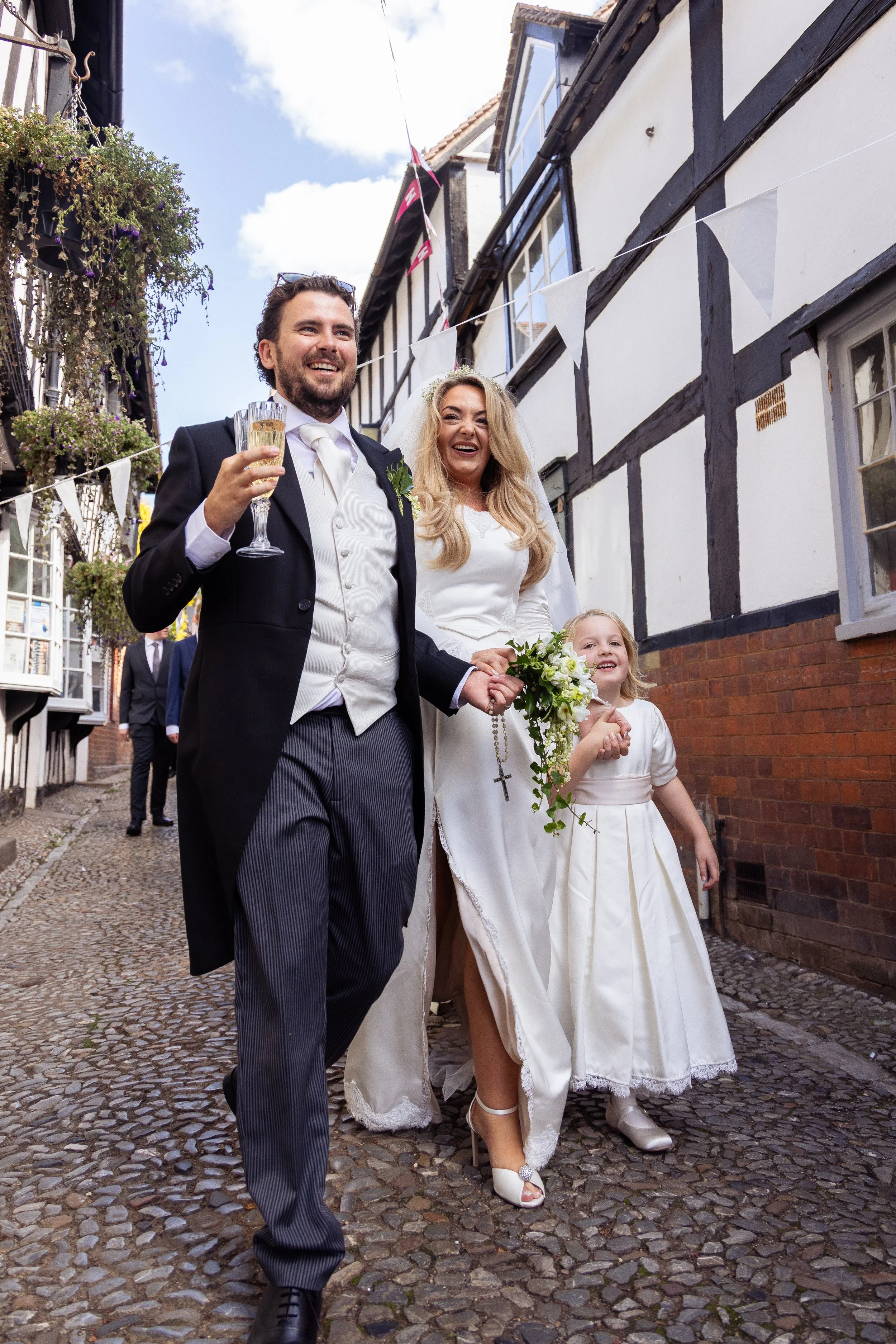 A happy groom and bride walking down a cobblestone street holding hands, accompanied by a young girl holding a bouquet, on a sunny day with decorative bunting hanging between buildings.