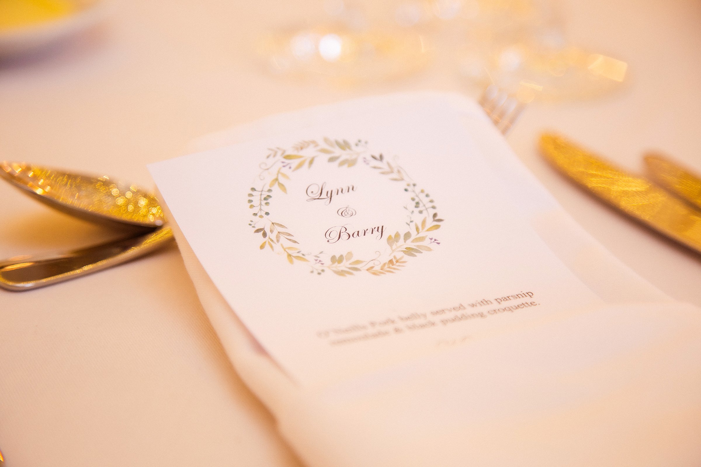 Close-up of an elegant wedding invitation with gold accents, featuring the names 'Lynn & Barry' surrounded by a decorative wreath design, placed on a soft pink surface with gold utensils partially visible.