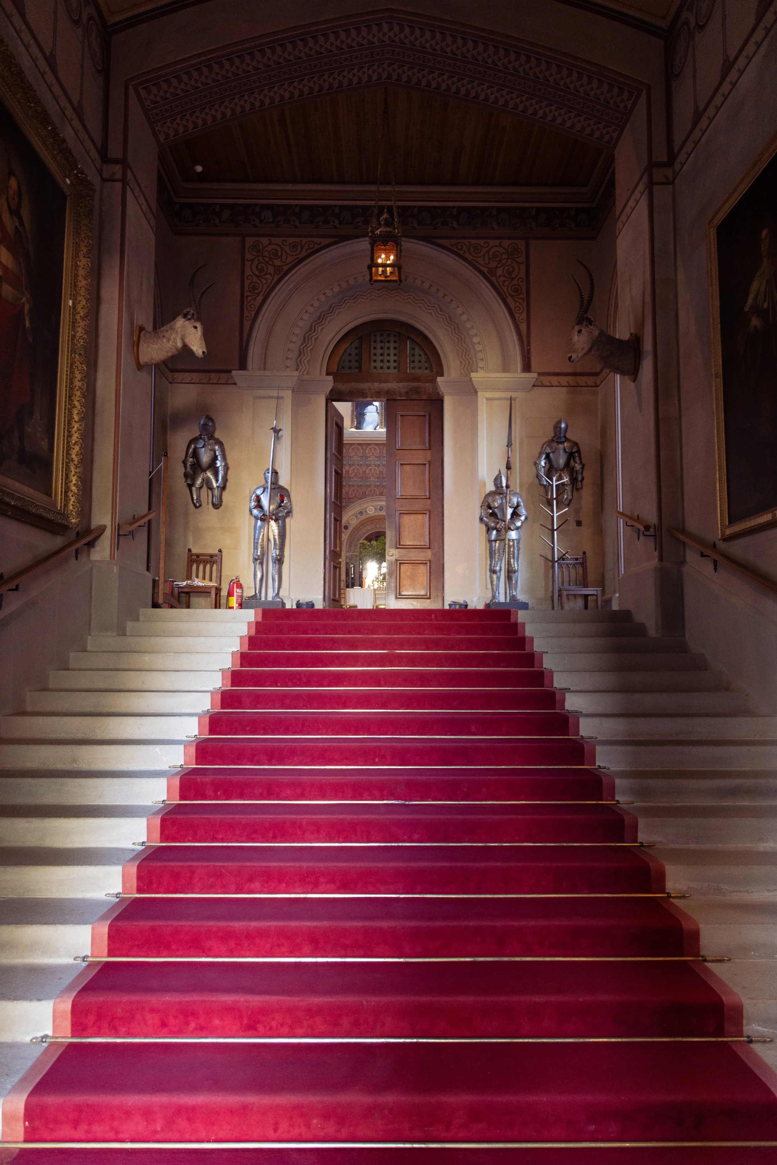 Grand staircase with red carpet runner, ornate walls, and animal head and knight armor decor in a historic hall.