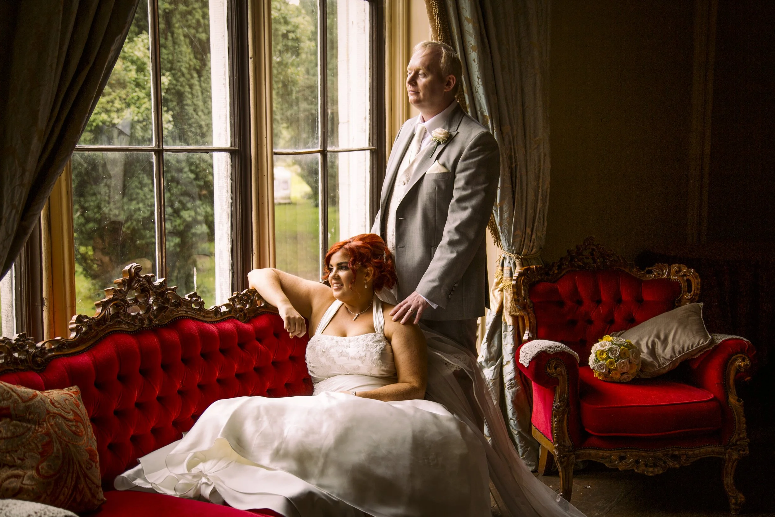 A man in a light gray suit standing behind a seated woman in a white wedding dress, both inside a room with ornate furniture and large windows, during daytime.