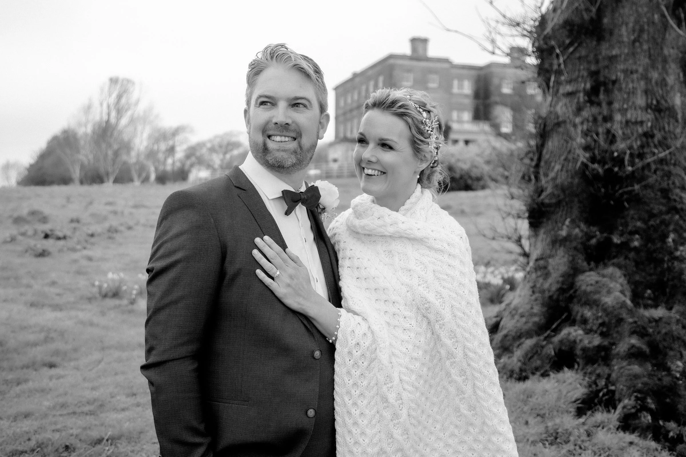 A black and white photo of a smiling bride and groom standing outdoors with a large house and trees in the background. The groom is dressed in a suit with a bow tie, and the bride is wearing a knitted shawl with a floral hairpiece.