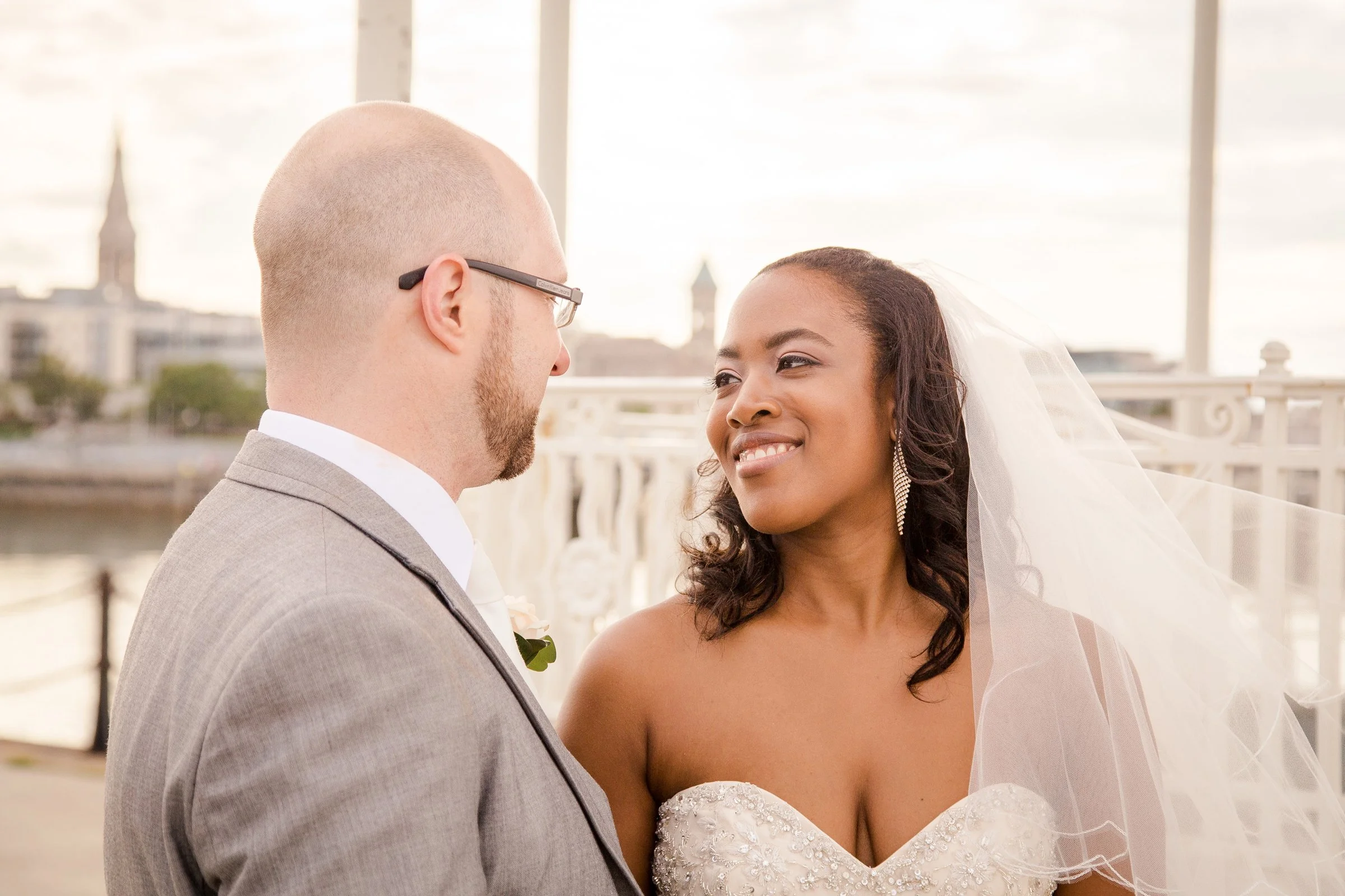A couple getting married outdoors on a bridge, with the groom in a gray suit and the bride in a white wedding dress and veil, gazing at each other lovingly.
