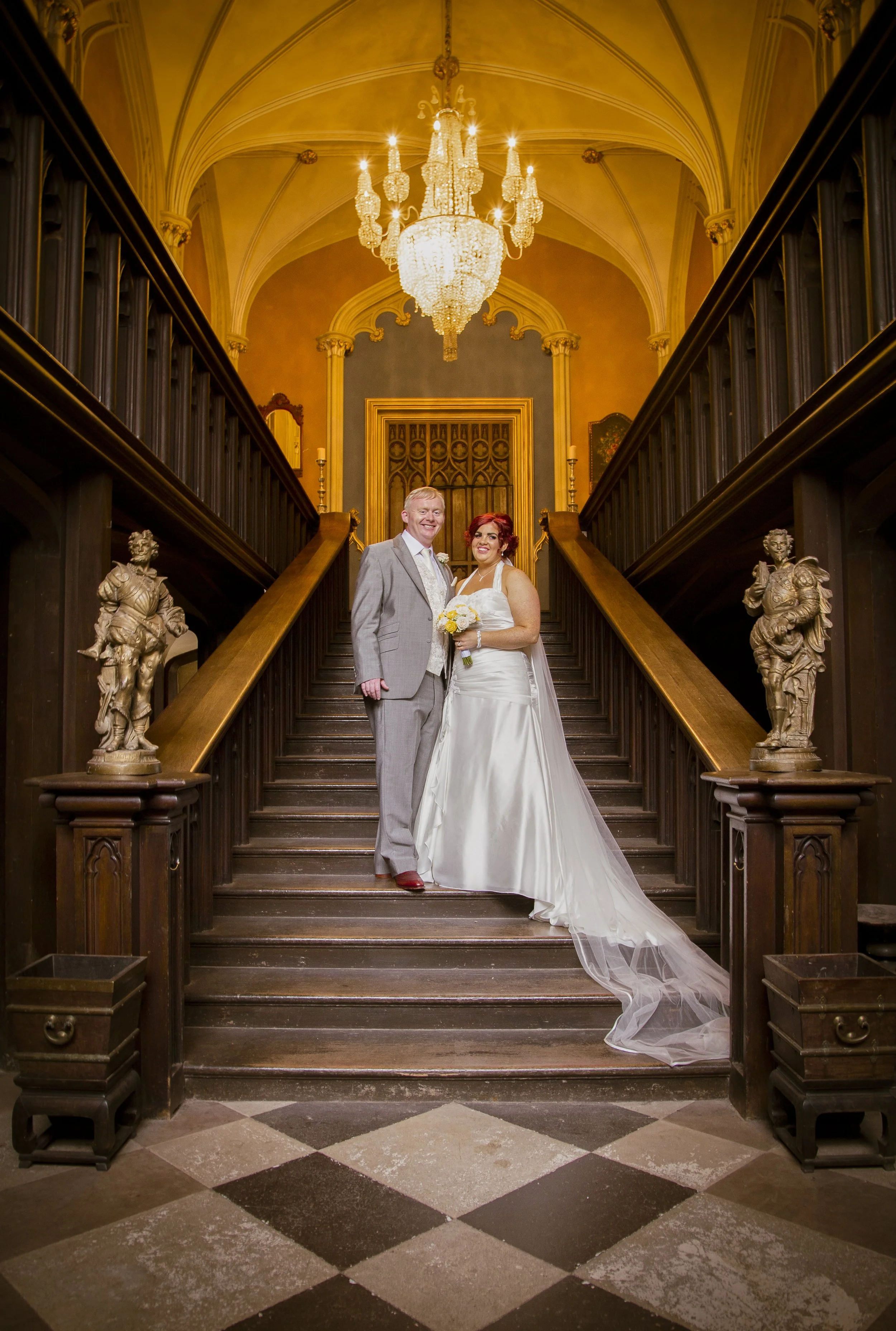 A bride and groom standing on a staircase inside a grand, ornate building with a large chandelier hanging above them. The couple is smiling, dressed in wedding attire, and surrounded by decorative statues on the staircase.
