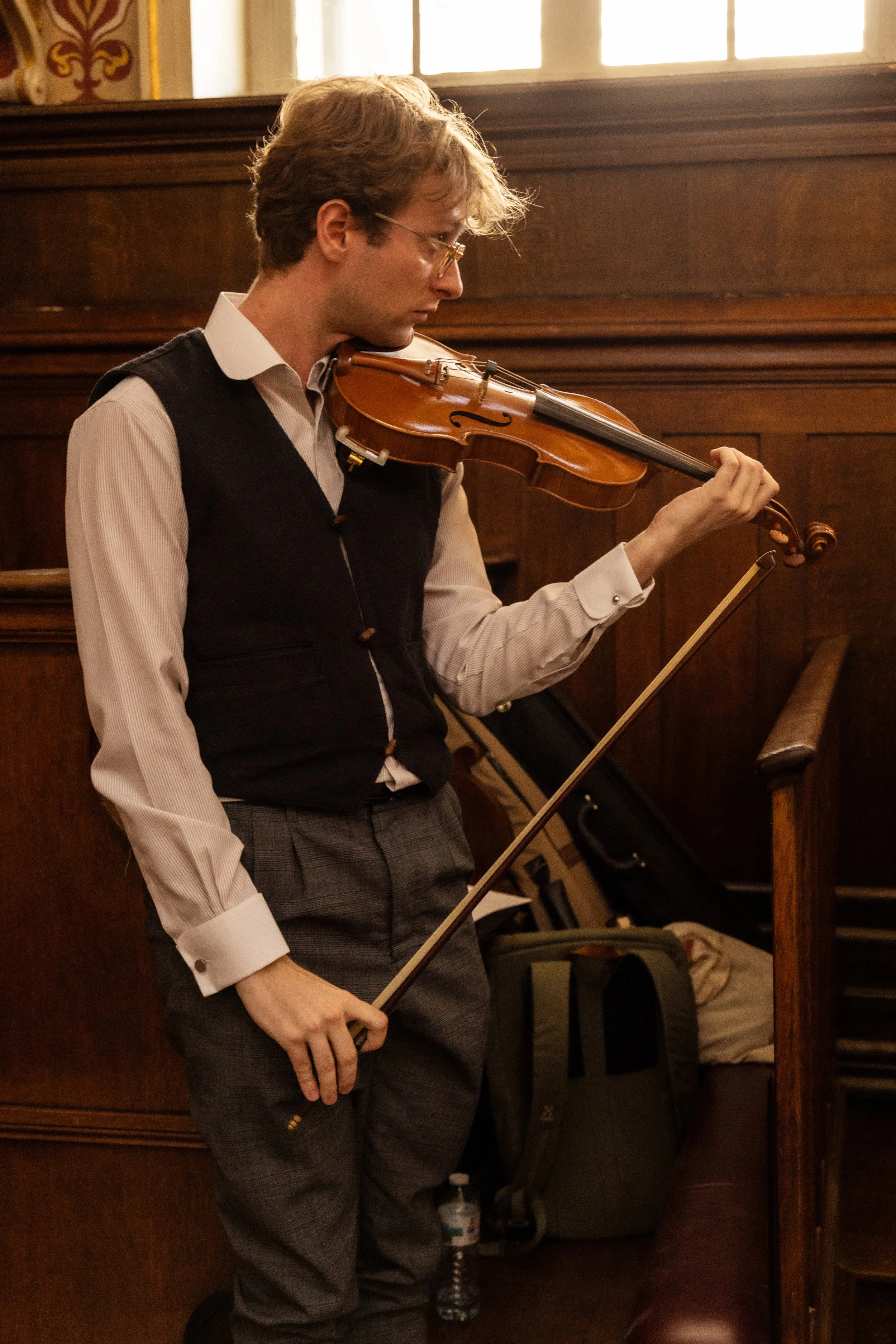A young man with light brown hair and glasses plays the violin in a room with wooden paneling and a window in the background.