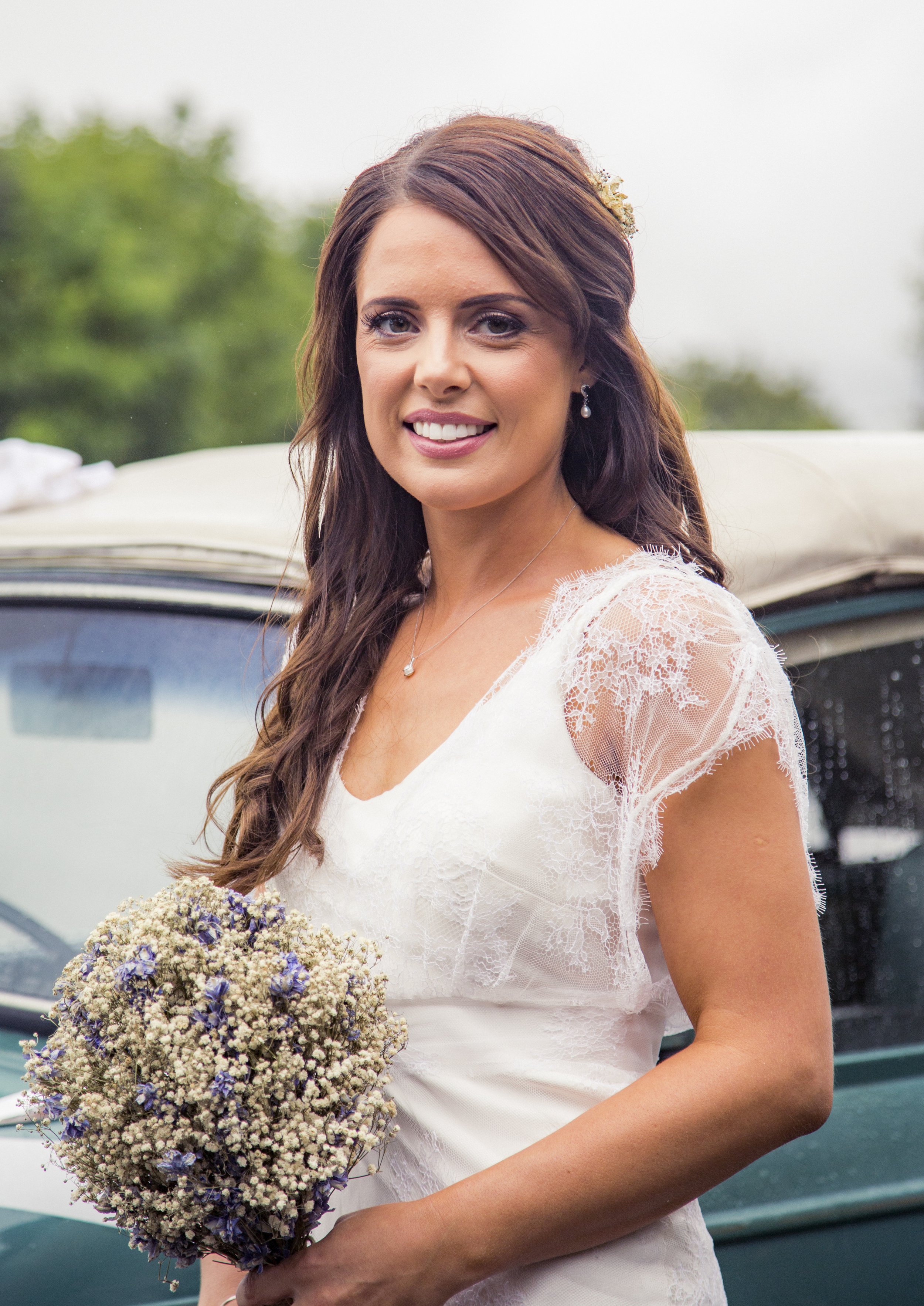 A woman in a wedding dress holding a bouquet of flowers stands outdoors near a vintage car, smiling at the camera.