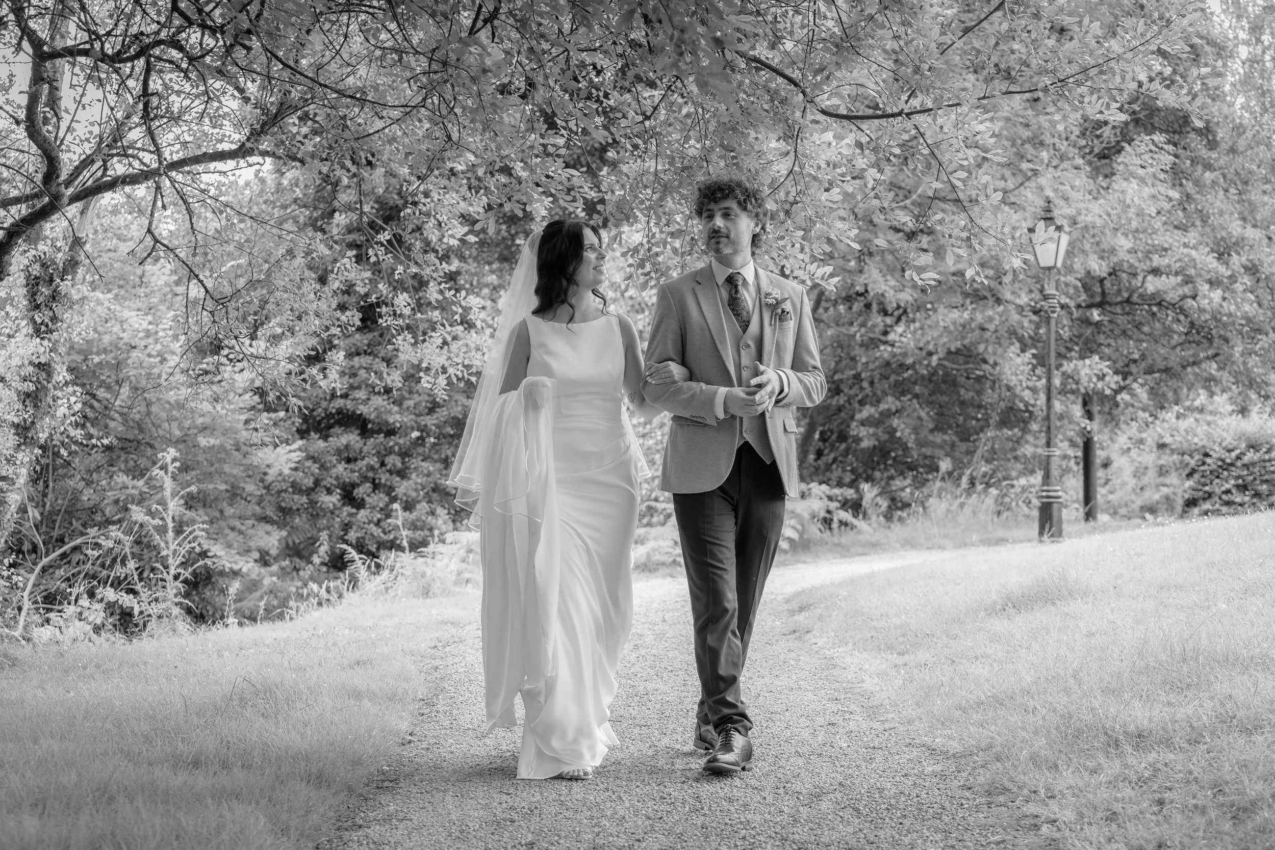 A black-and-white photo of a bride and groom walking outdoors under a large tree. The bride is wearing a white wedding dress and veil, and the groom is dressed in a suit. They are holding onto each other, and there is a lamppost behind them.