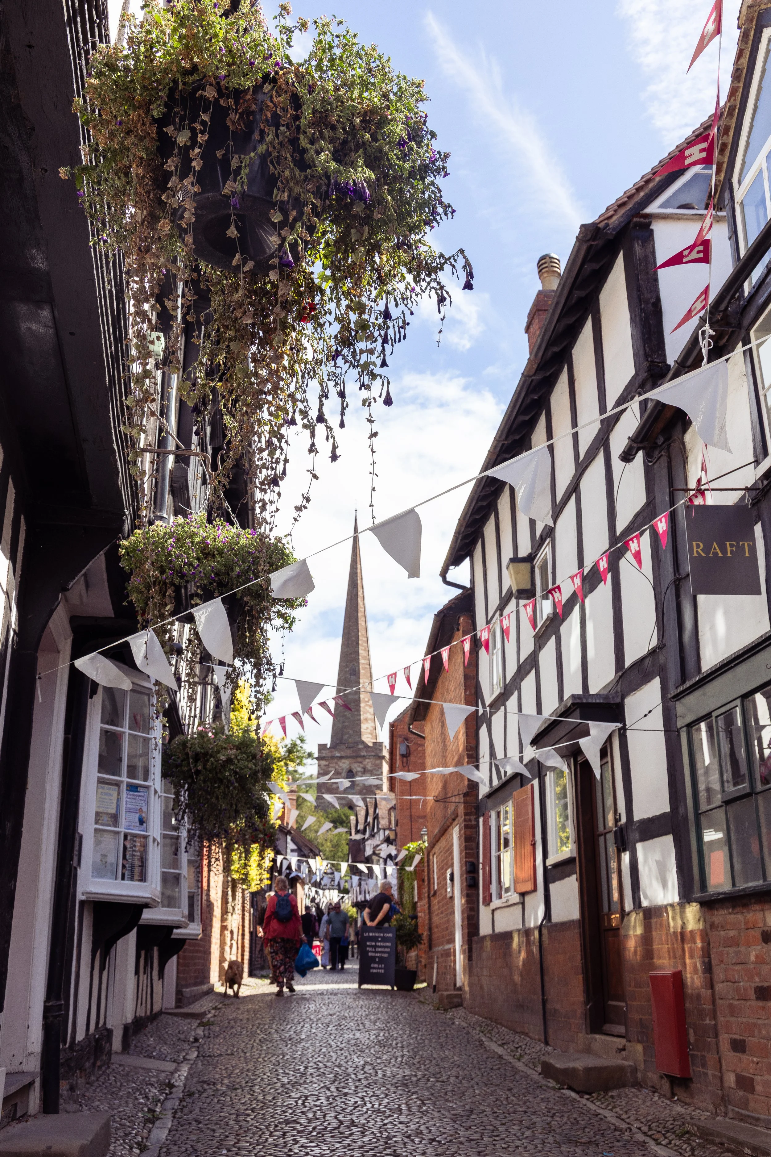 A narrow cobblestone street in a historic area with hanging flower baskets, bunting, and people walking. Buildings have traditional timber framing and brickwork, with a church steeple visible in the background.