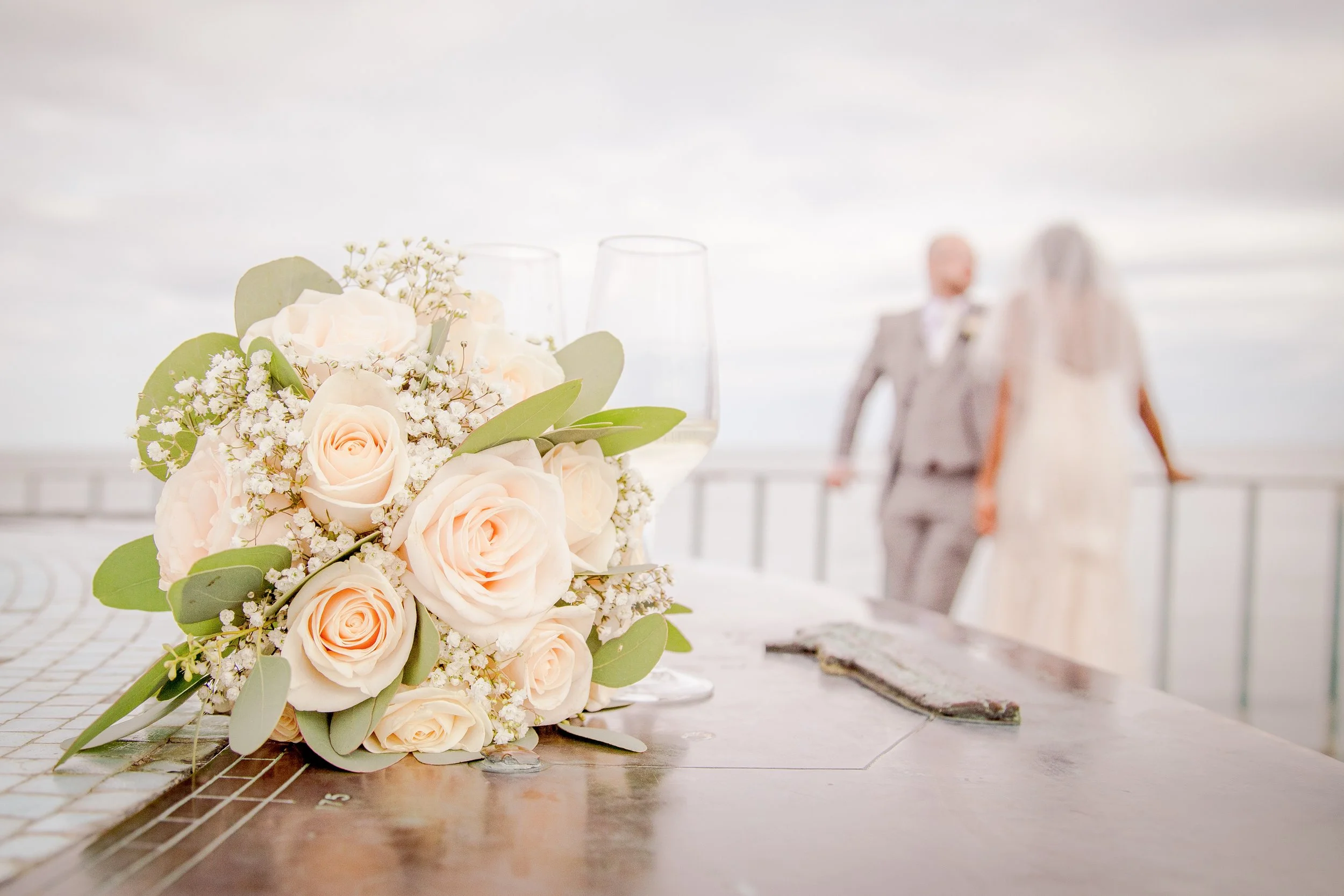 Close-up of a white and blush pink floral bouquet with baby's breath on a tiled floor at a wedding venue, with a blurred bride and groom in the background.