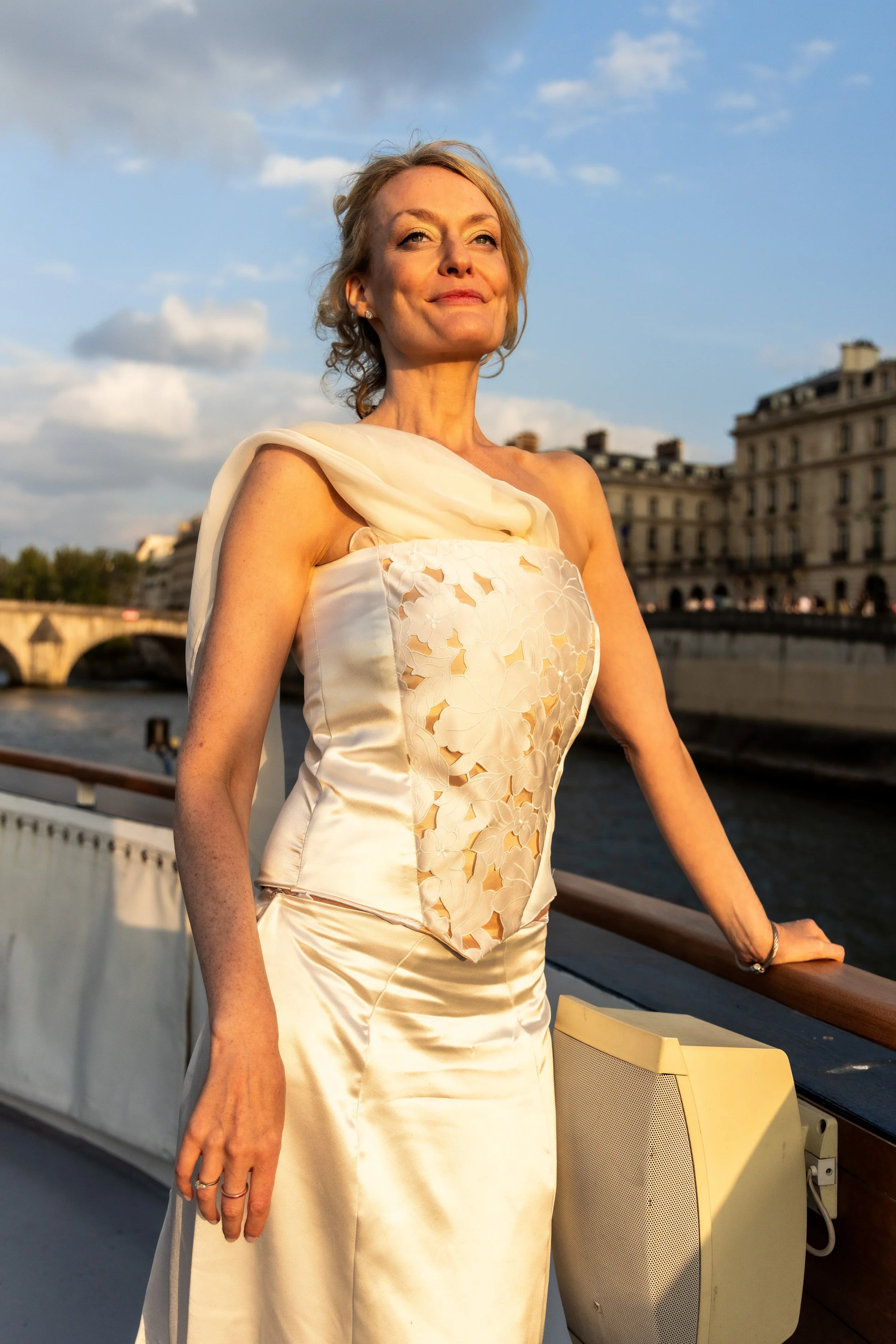A woman in a white dress with floral embroidery standing on a boat deck, with the river and historic buildings in the background during sunset.