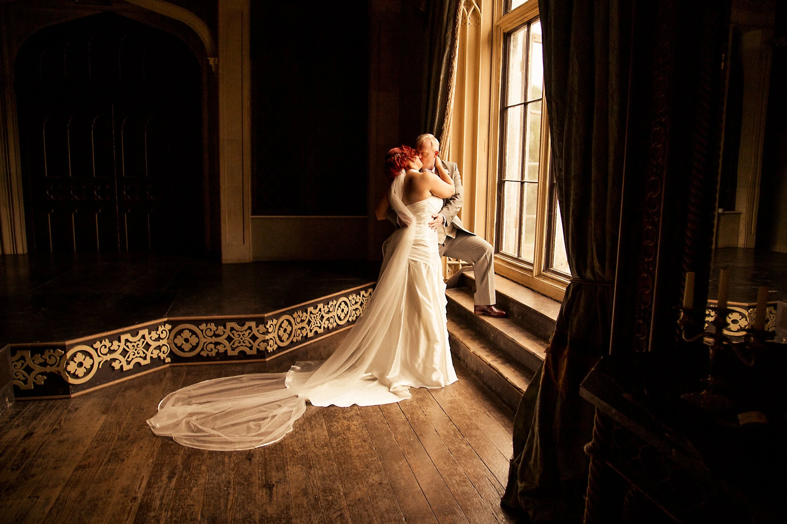 A bride and groom sharing a kiss by a large window, with the bride in a white wedding dress and the groom in a gray suit, in a dimly lit room with dark wood floors and ornate paneling.