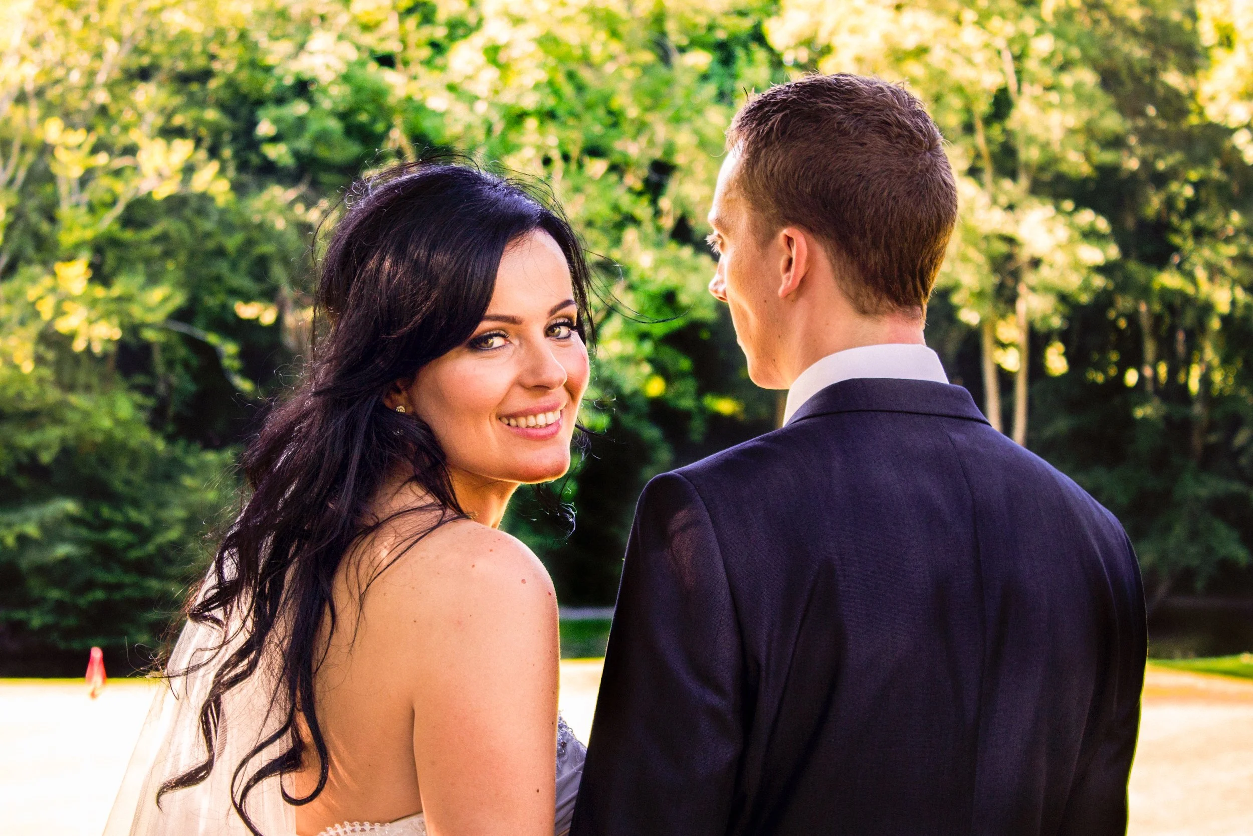A smiling woman with dark hair and a man with short hair in a suit outdoors among green trees.