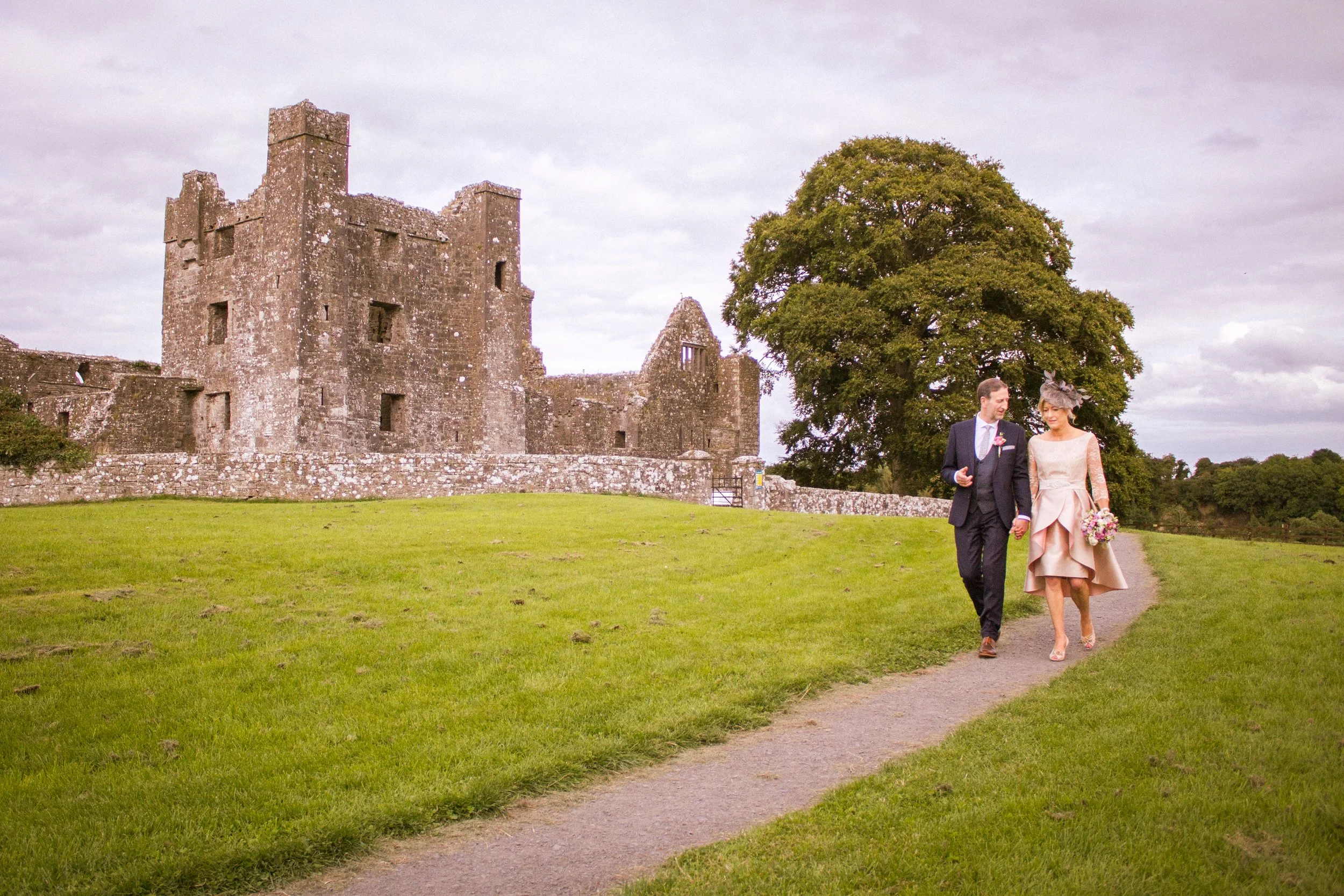 A couple dressed in wedding attire walking hand in hand on a dirt path in front of an old stone castle and large green tree.
