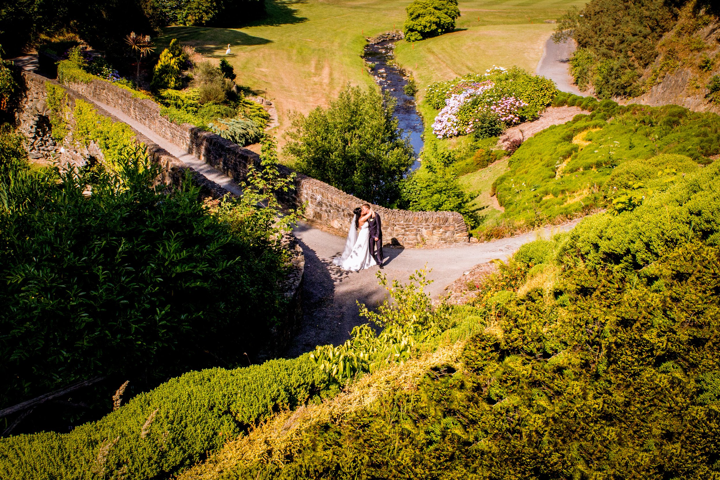 A bride and groom kissing in a lush garden with a stone wall, a small stream, trees, flowers, and a grassy hill.