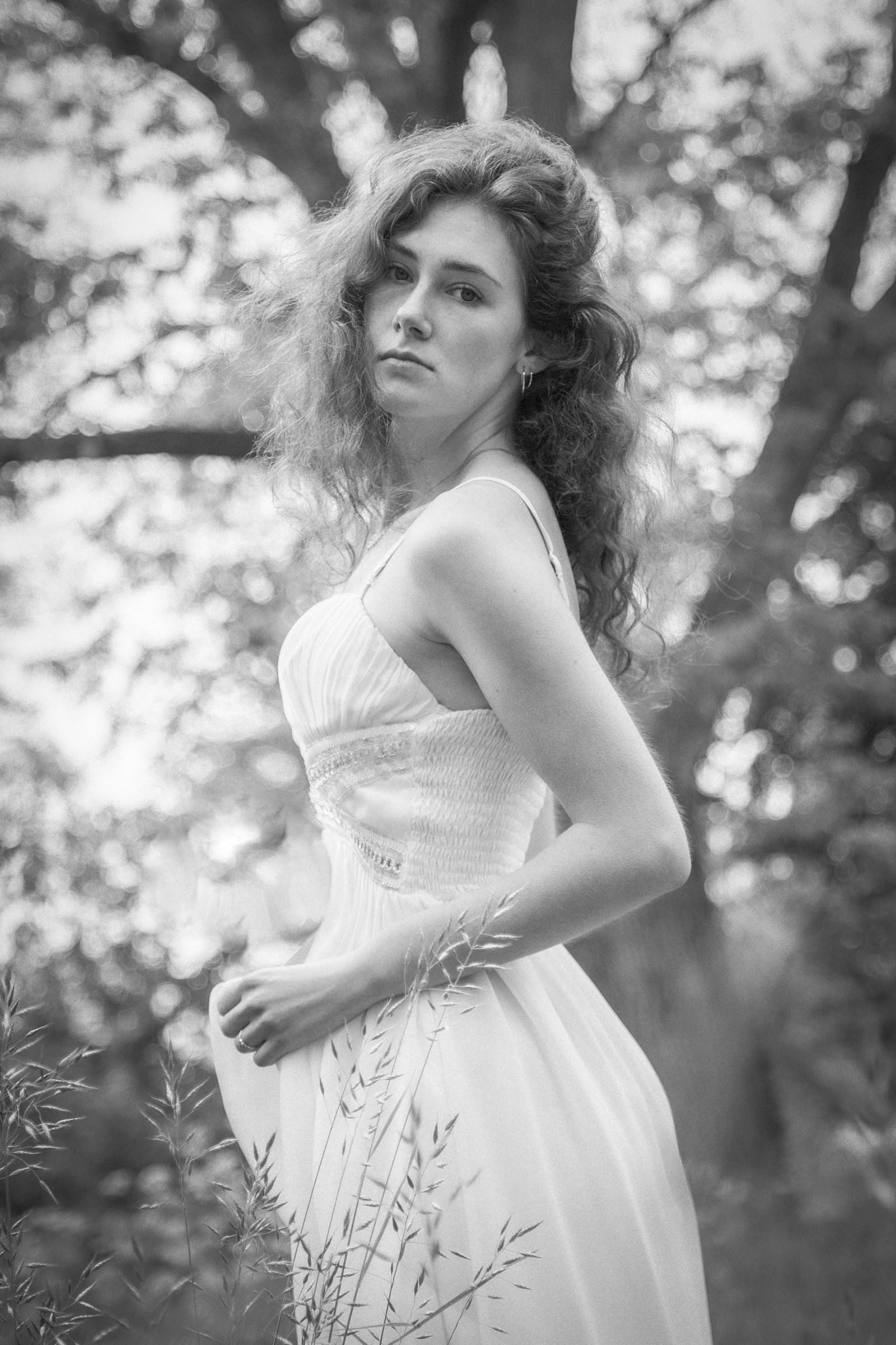 A black-and-white photo of a young woman with wavy hair, wearing a light-colored dress, standing outdoors among trees and grass.