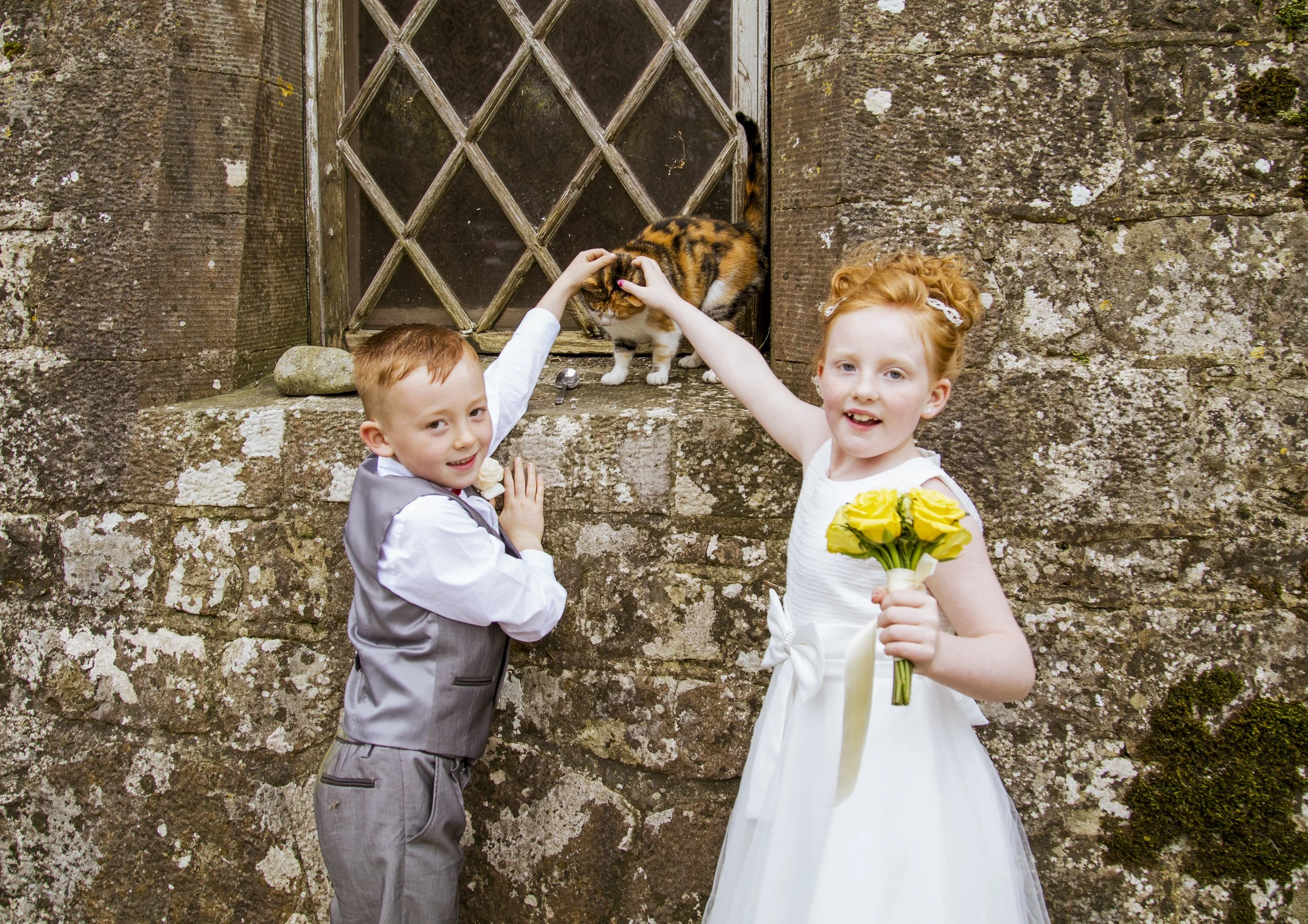 Two children, a young boy and girl, are dressed in formal wedding attire and holding yellow flowers. They are standing in front of a stone wall with a small window, with the girl holding a bouquet of yellow roses and the boy adjusting his vest. A cal