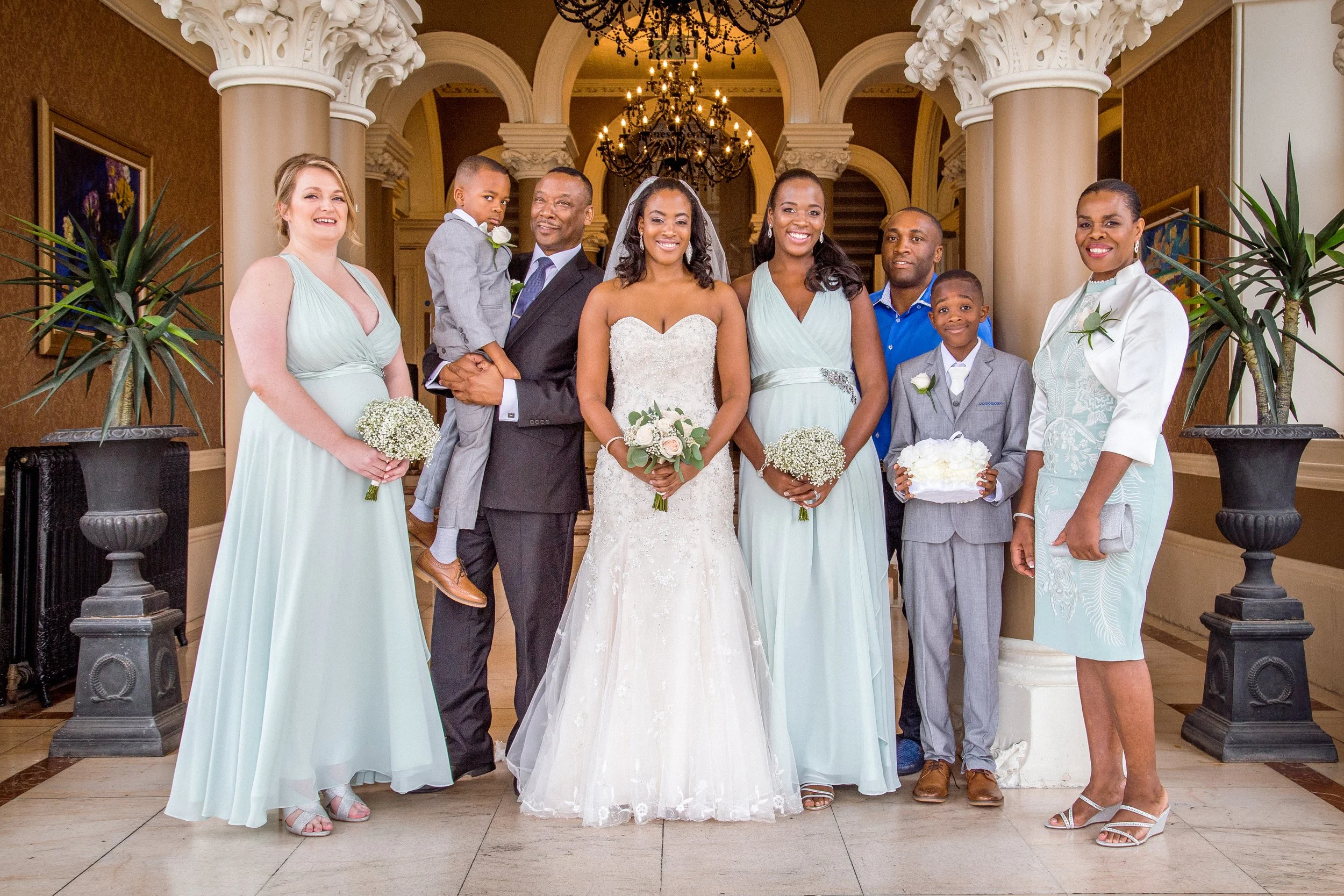 A group of nine people, including a bride in a white wedding gown, posed for a photo in an elegant indoor setting with chandeliers and classical columns. The group includes men, women, and children dressed in formal attire, some holding bouquets of f
