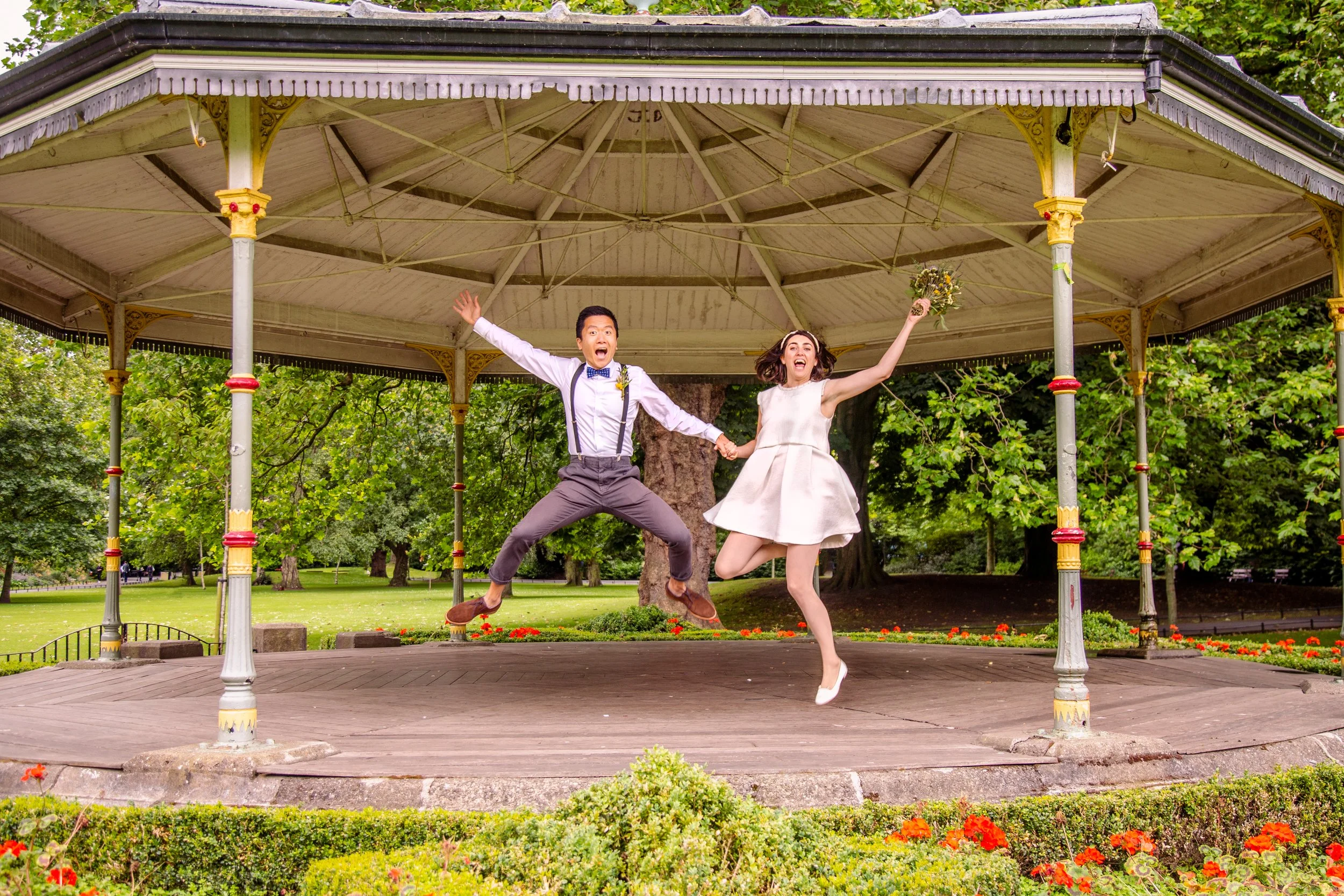 A joyful couple jumping in the air on a vintage gazebo in a park, holding hands with trees and flowers in the background.
