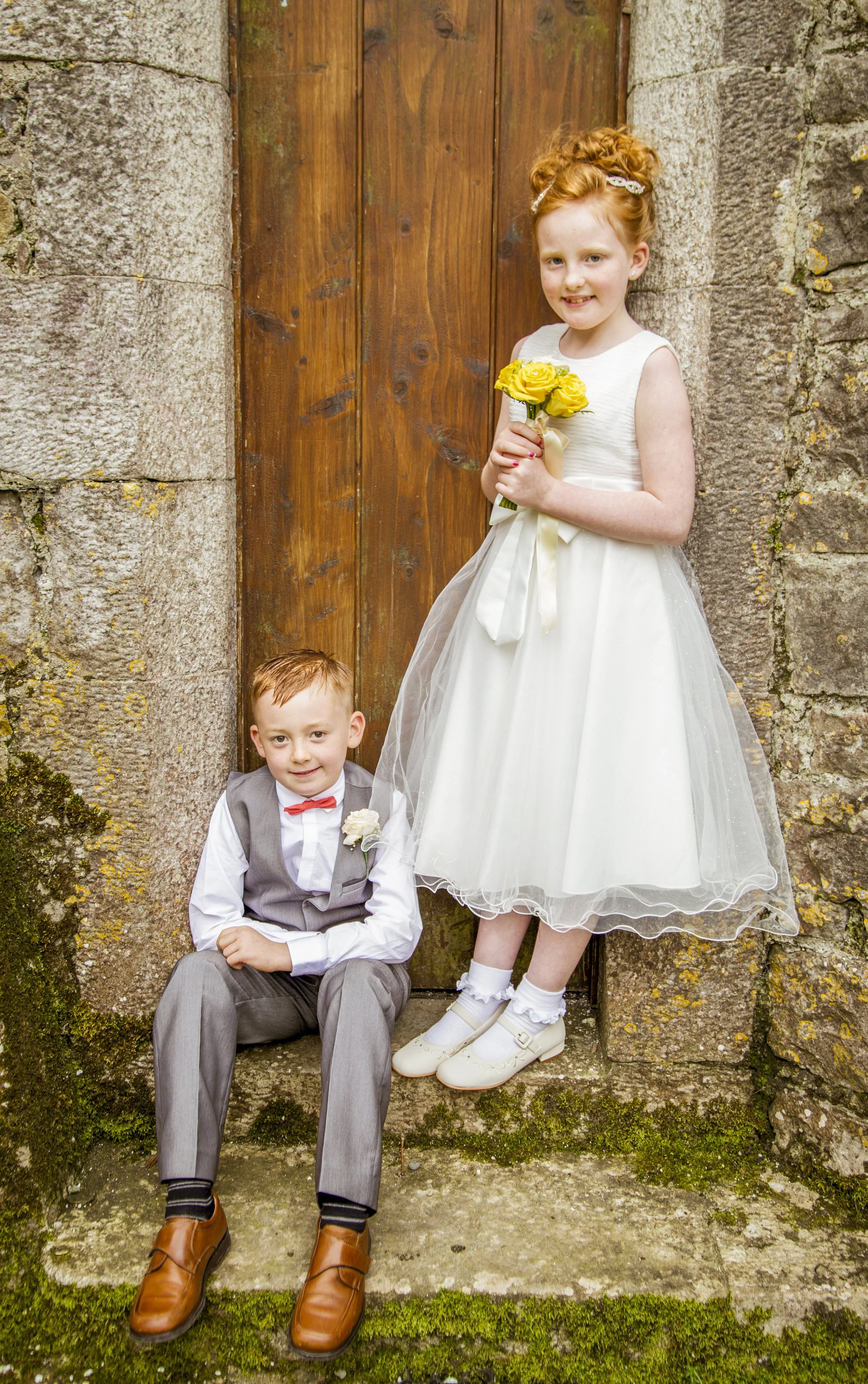 A young girl in a white dress holding a yellow flower bouquet standing beside a young boy in a gray vest and bow tie sitting on steps in front of a wooden door.