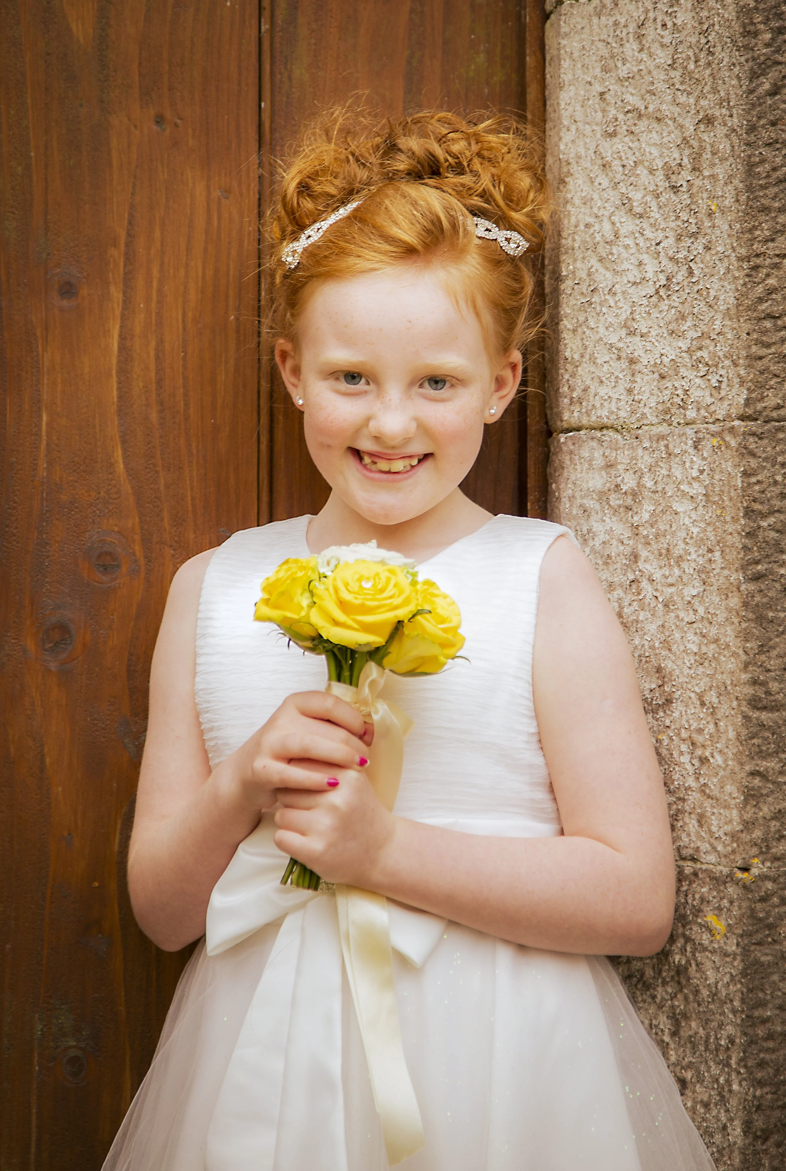 A young girl with red hair styled in an updo, wearing a white dress with a satin ribbon, holding a bouquet of yellow roses, standing against a wooden and stone background.