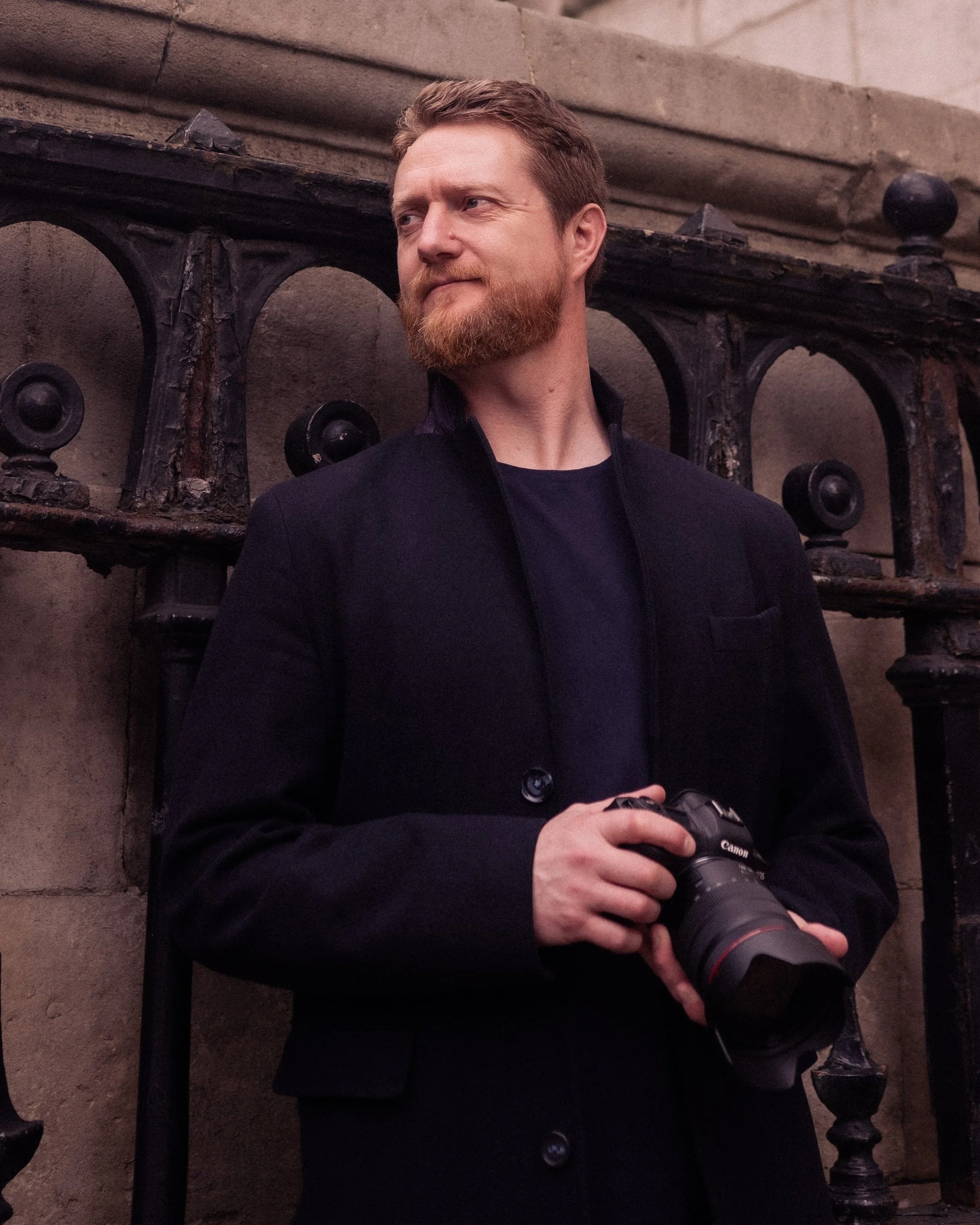 A man with a red beard and brown hair wearing a black jacket, holding a professional camera with a long lens, standing in front of a stone wall with a metal railing.