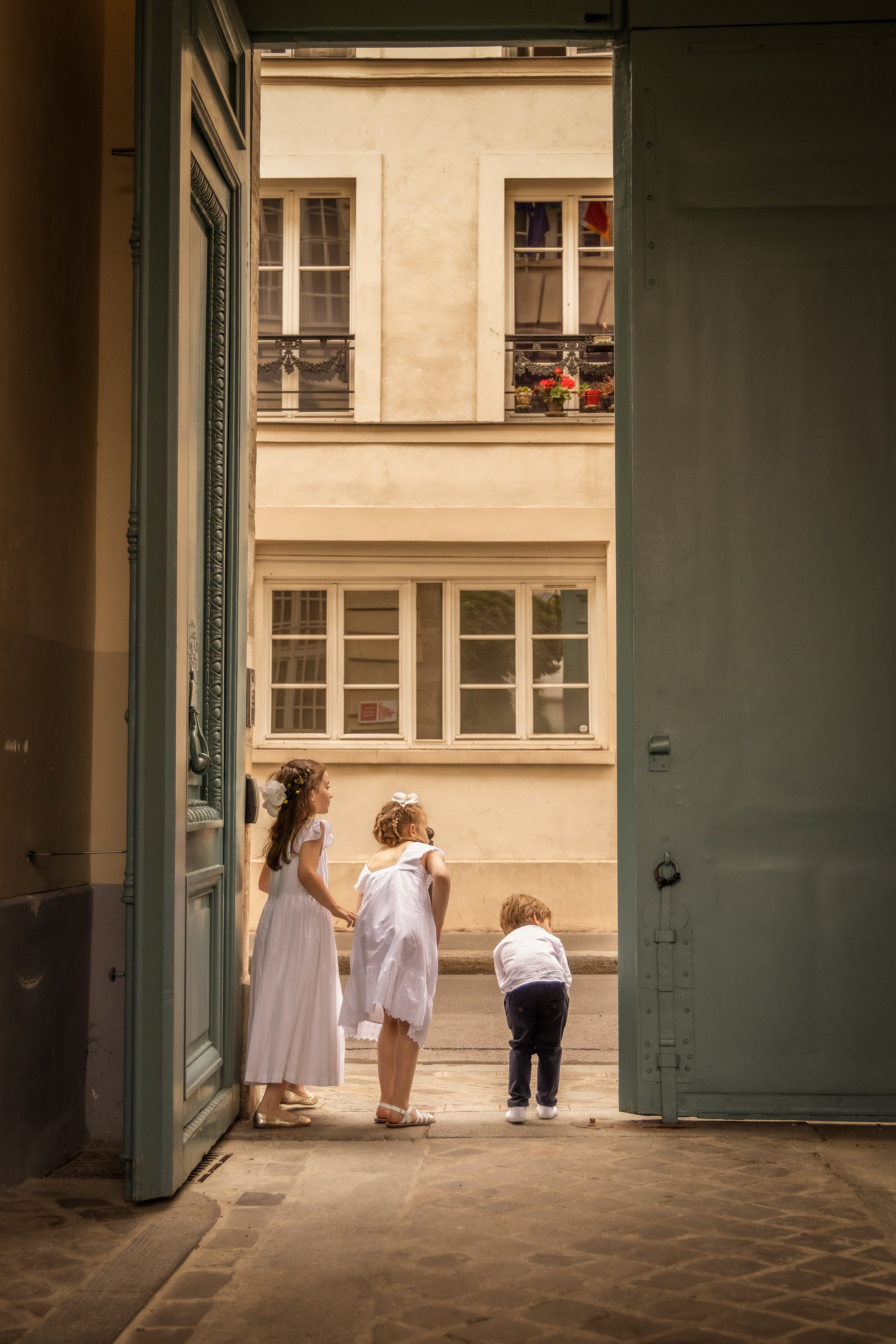 Children dressed in white clothes standing and kneeling outside a building, viewed through an open door with ornate framing, in a European-style street setting.
