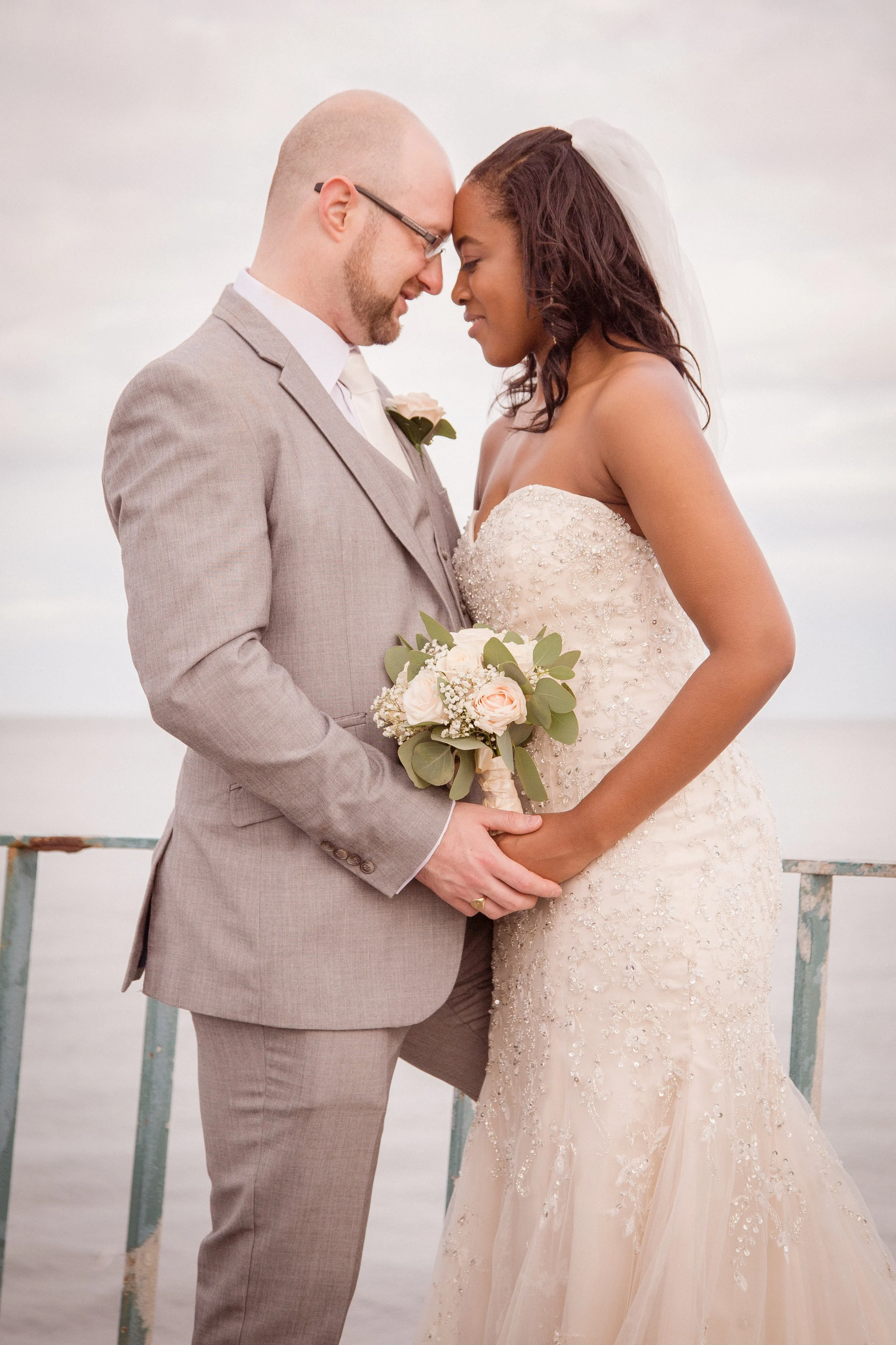 A newly married couple stands close together with foreheads touching. The man is wearing a light gray suit and glasses, while the woman is in a strapless, beaded wedding gown and veil. She holds a bouquet of white and pink flowers. They are outdoors 