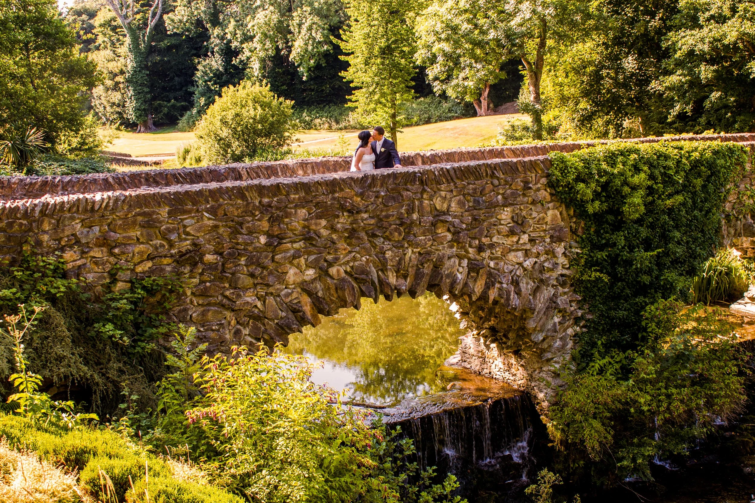 A stone bridge over a small waterfall in a lush, green park with large trees and foliage.