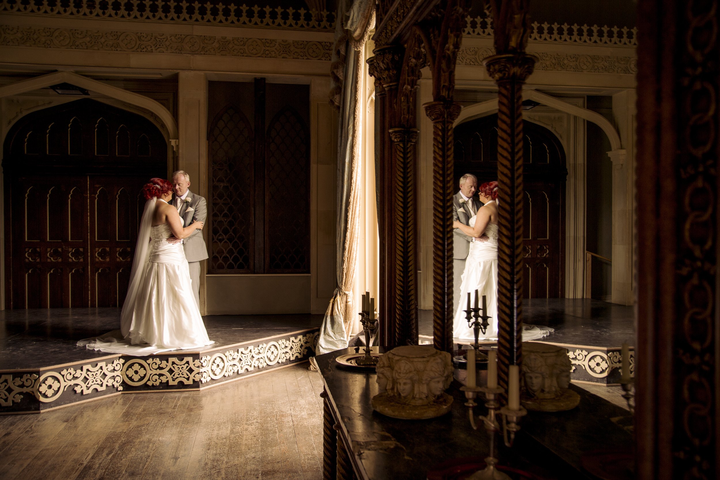 A bride and groom standing on a small stage in a dimly lit elegant room, holding hands and looking at each other. The bride is wearing a white strapless gown with a veil, and the groom is in a light gray suit with a bow tie. The room features ornate 