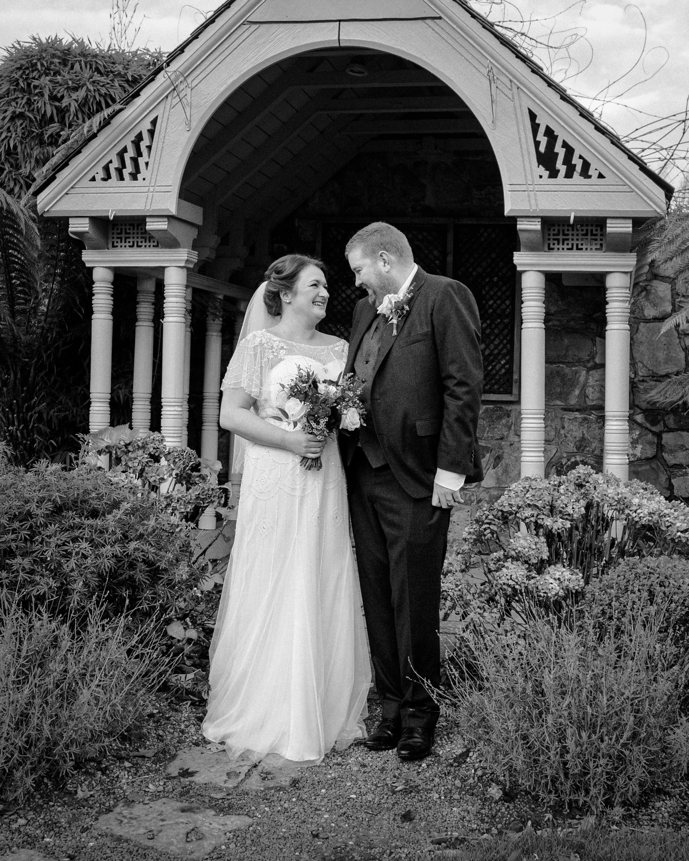 Black and white photo of a bride and groom holding hands under a decorative outdoor structure, smiling at each other, surrounded by plants and flowers.