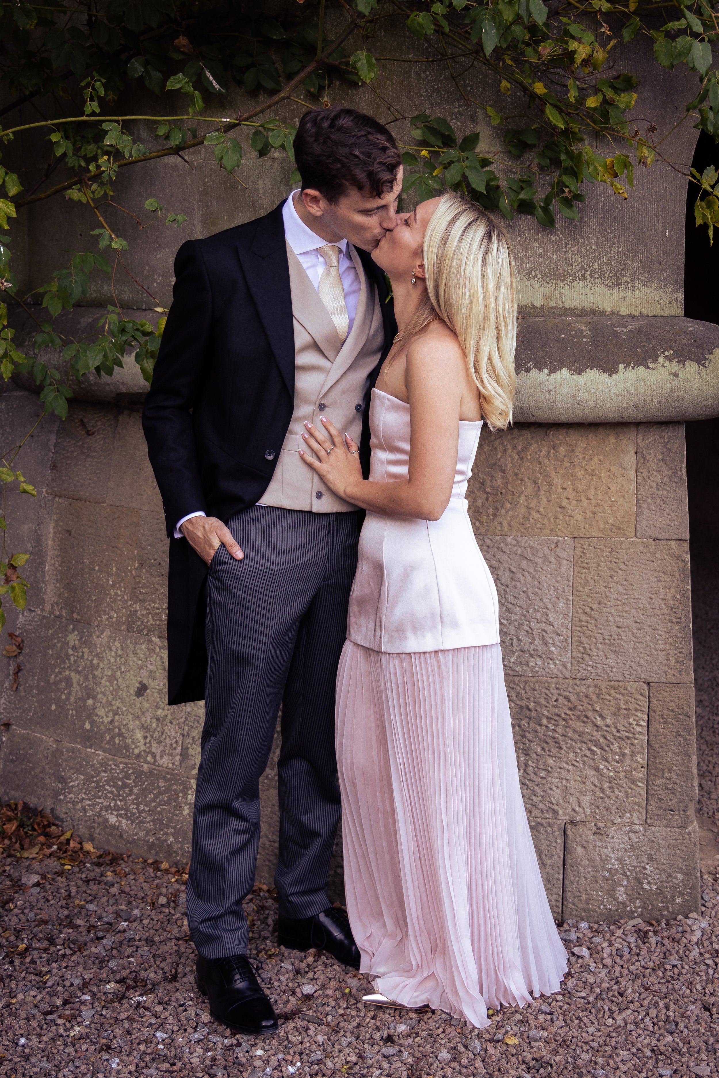 A newly married couple sharing a kiss outside in front of a stone wall covered in green ivy, with the bride wearing a strapless pink gown and the groom in a black suit with beige vest and white tie.