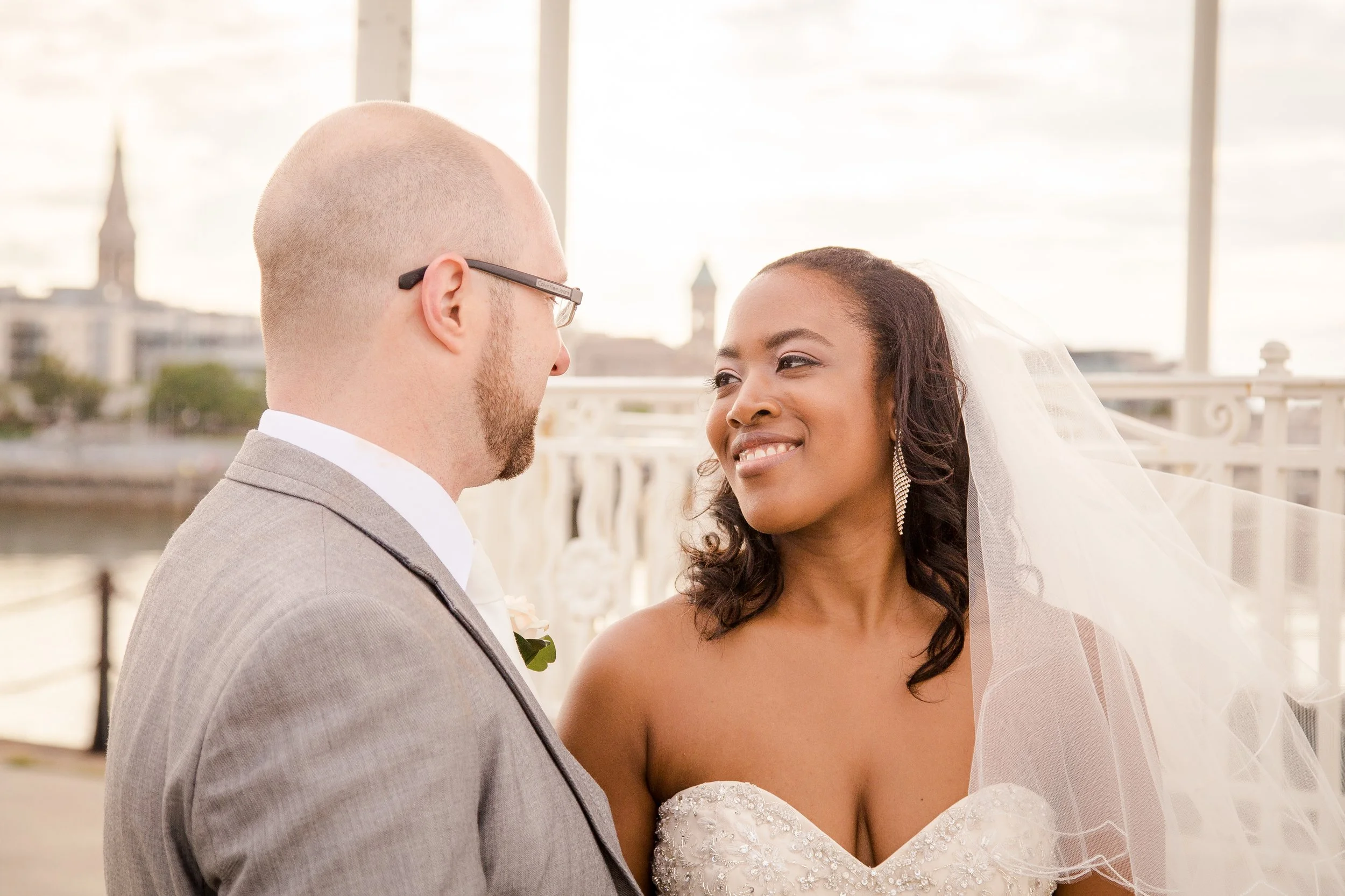 A newlywed couple faces each other on a bridge with a city skyline in the background, bride smiling and bridegroom looking at her.