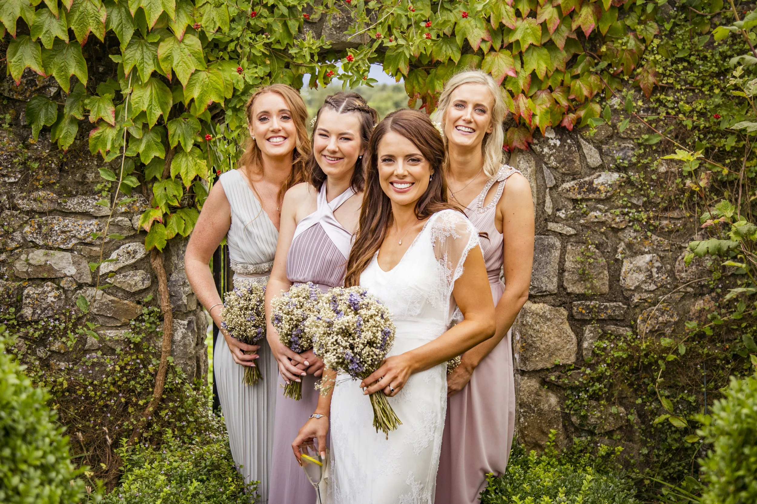A group of five women dressed in formal attire, smiling, standing outdoors in front of a stone and vine-covered wall during daytime.