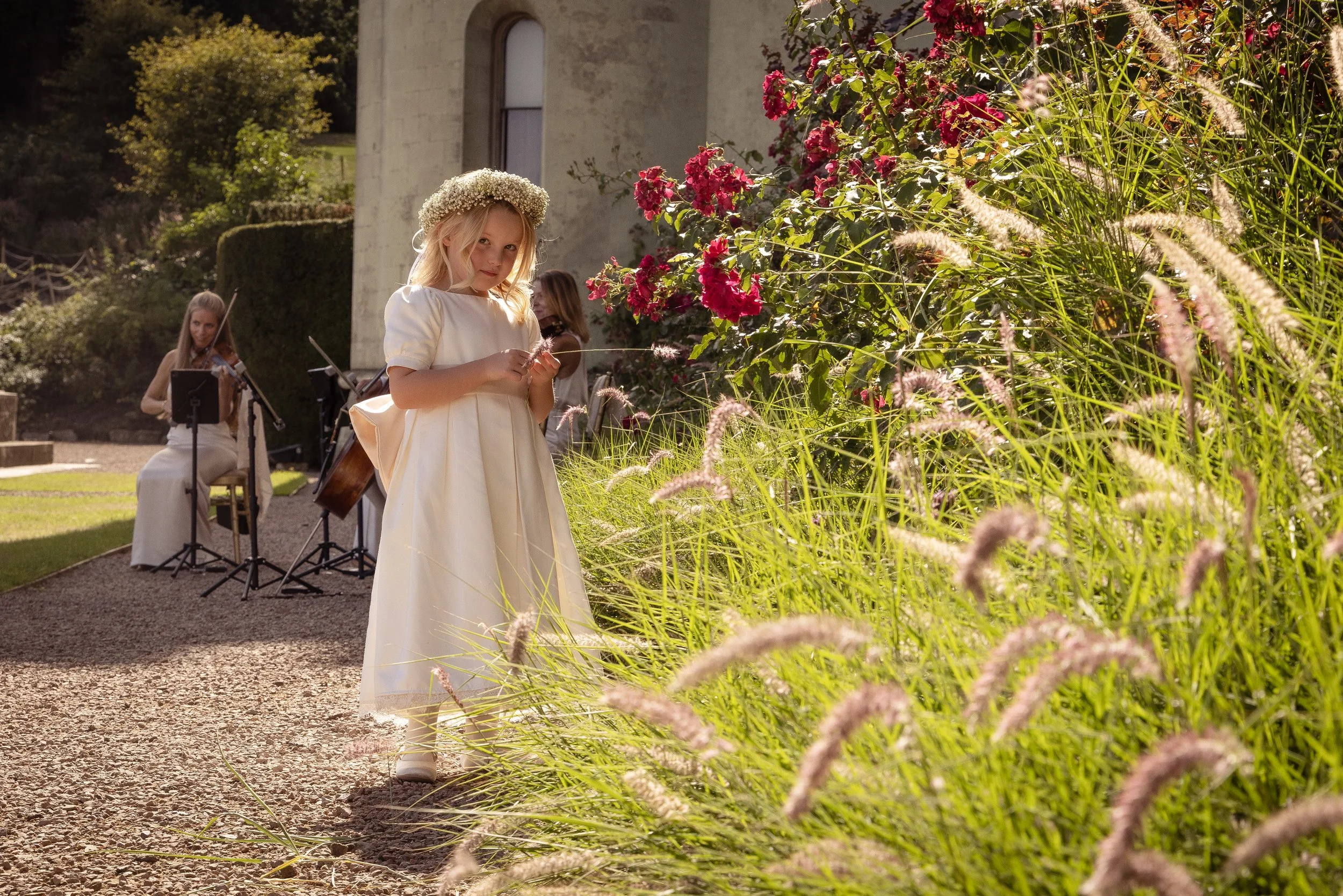 A young girl dressed in a white gown with a floral crown on her head standing in a garden surrounded by pink flowers, with musicians playing instruments in the background.
